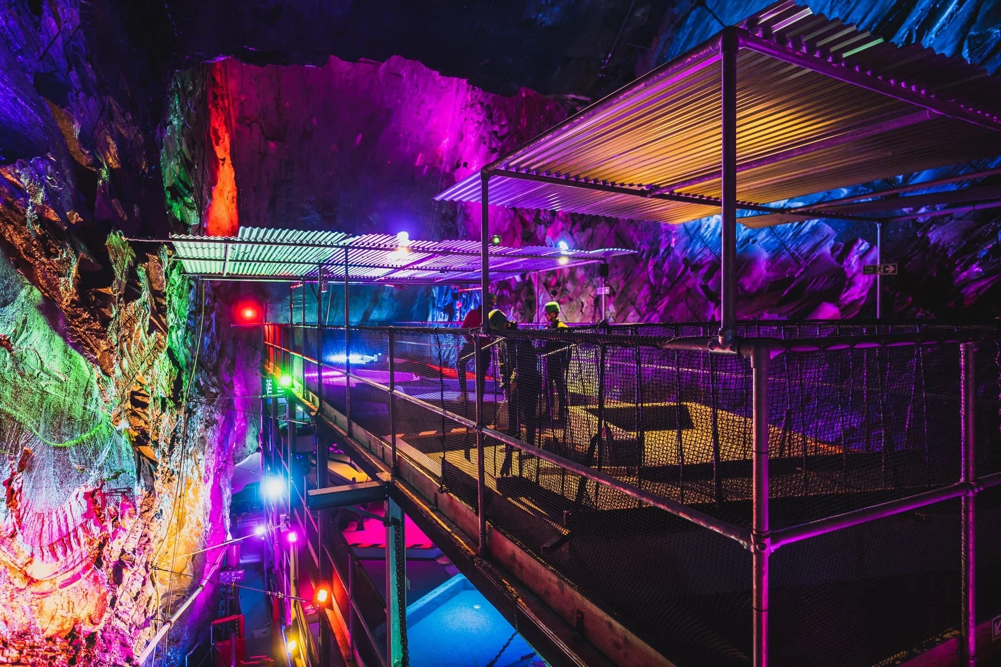 People standing on a walkway inside Zip World Llechwedd's Bounce Below, a colorful, illuminated cave with neon lights and rock formations.