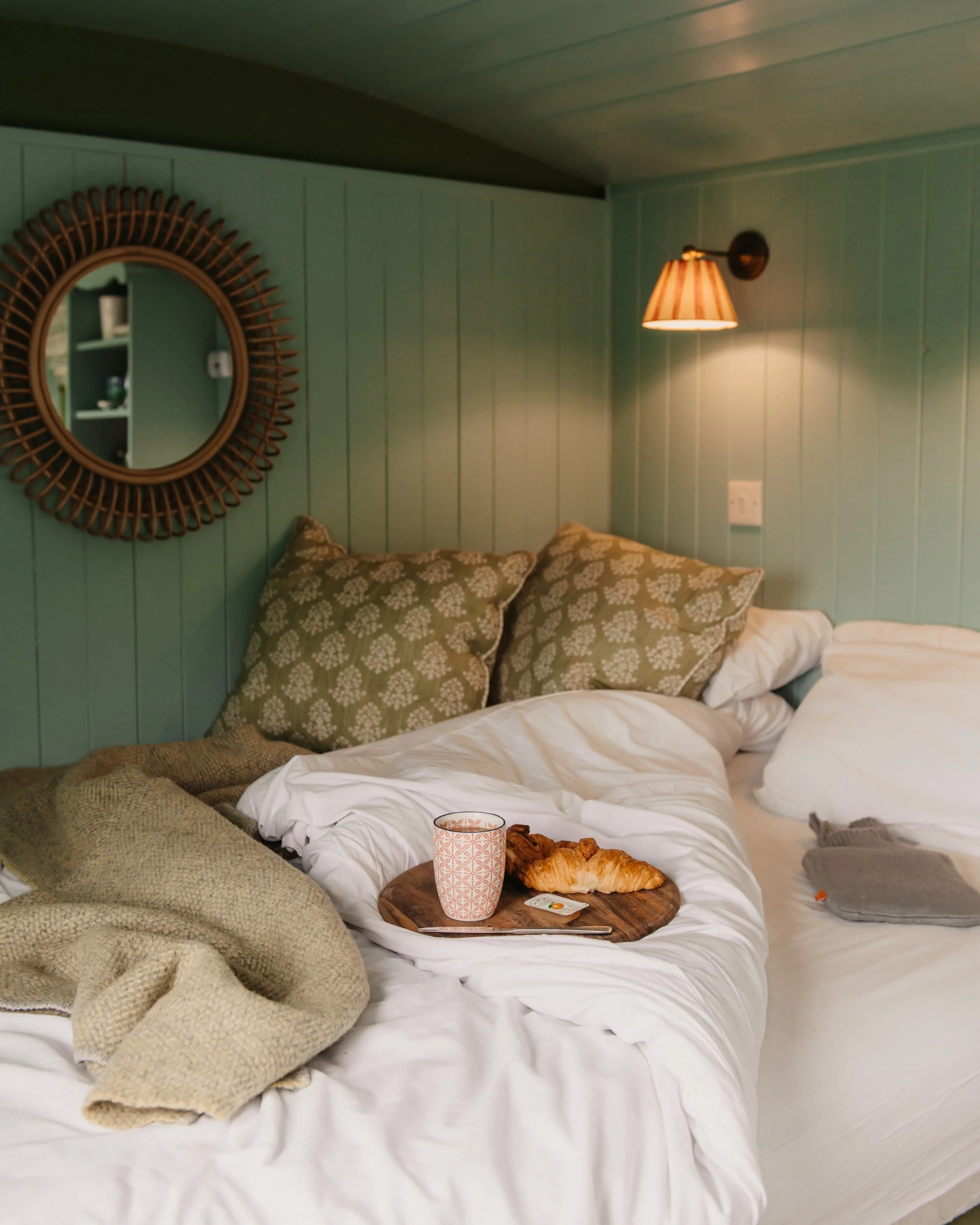 A cozy bed inside a Barlwyd Shepherd's Hut with white sheets and pillows, a tray with a croissant and a cup, and a blanket, set against a wooden paneled wall with a mirror and wall lamp.