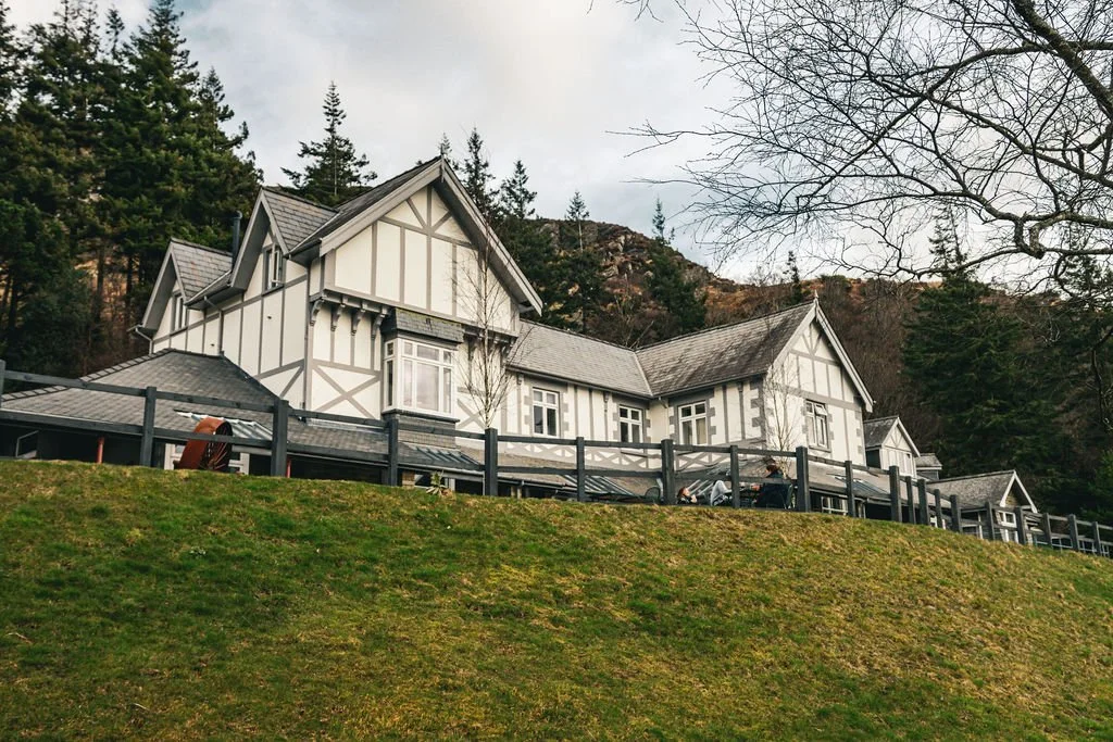 A large, Tudor-style house with white walls and dark wooden beams on a hillside, surrounded by trees and a grassy yard.