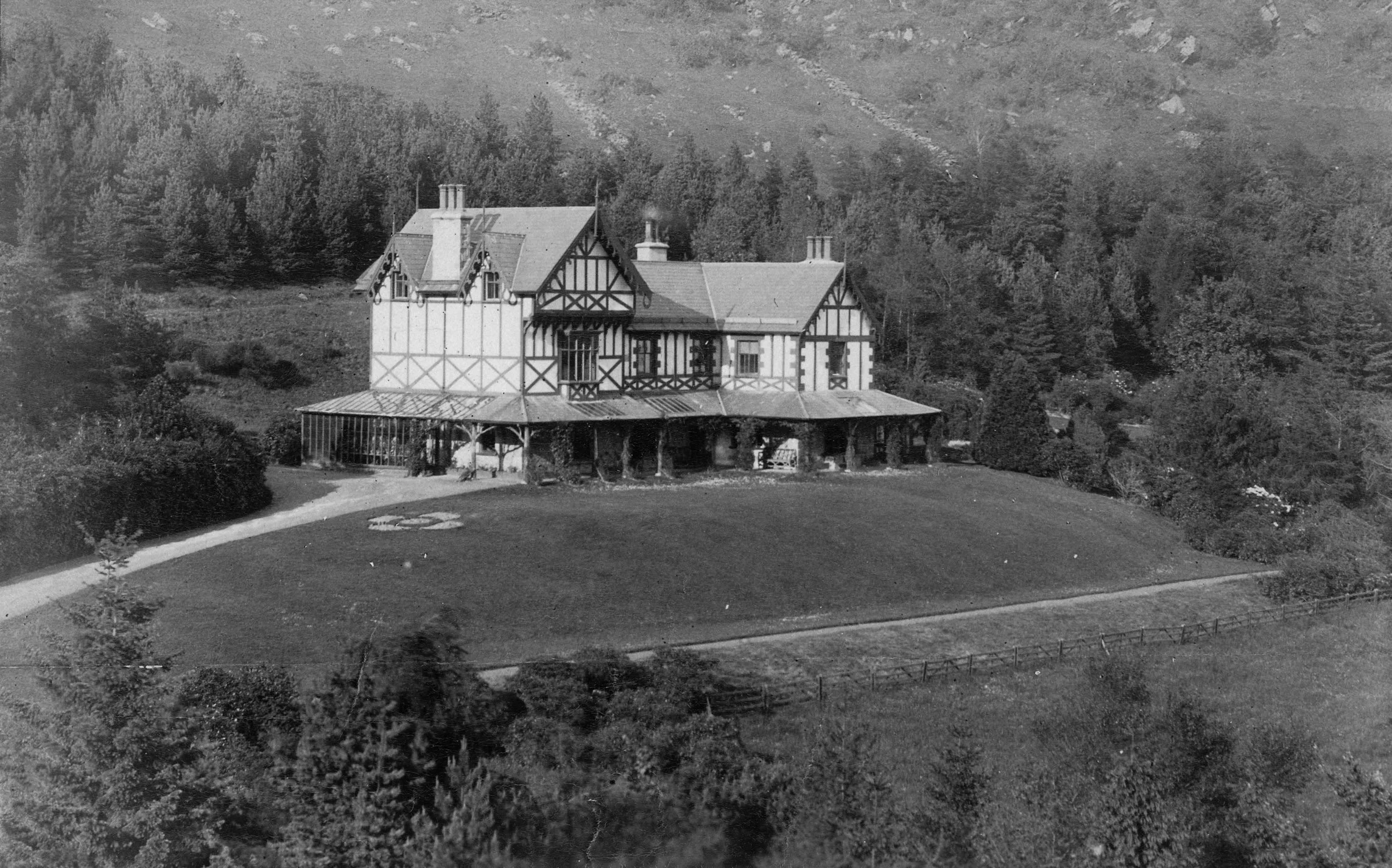 A black and white historical photo of Plas Weunydd. A large house with Tudor-style architecture, surrounded by trees, with a lawn and a path leading to the house in a mountainous area.