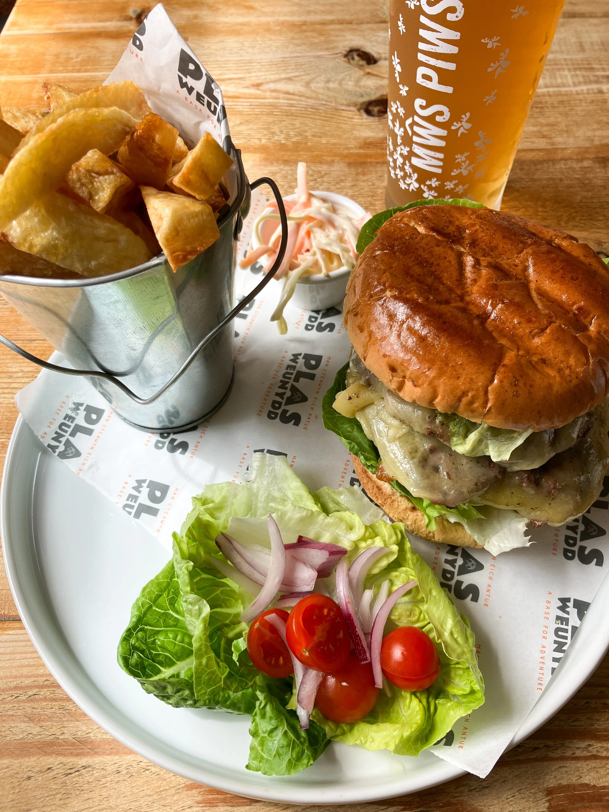 A cheeseburger with lettuce, pickles, and melted cheese on a toasted bun, served with a side salad of cherry tomatoes, red onion slices, and lettuce, alongside a small serving of coleslaw, a bucket of thick-cut fries, and a pint of beer with the logo 'Twin's Pines' on the glass.