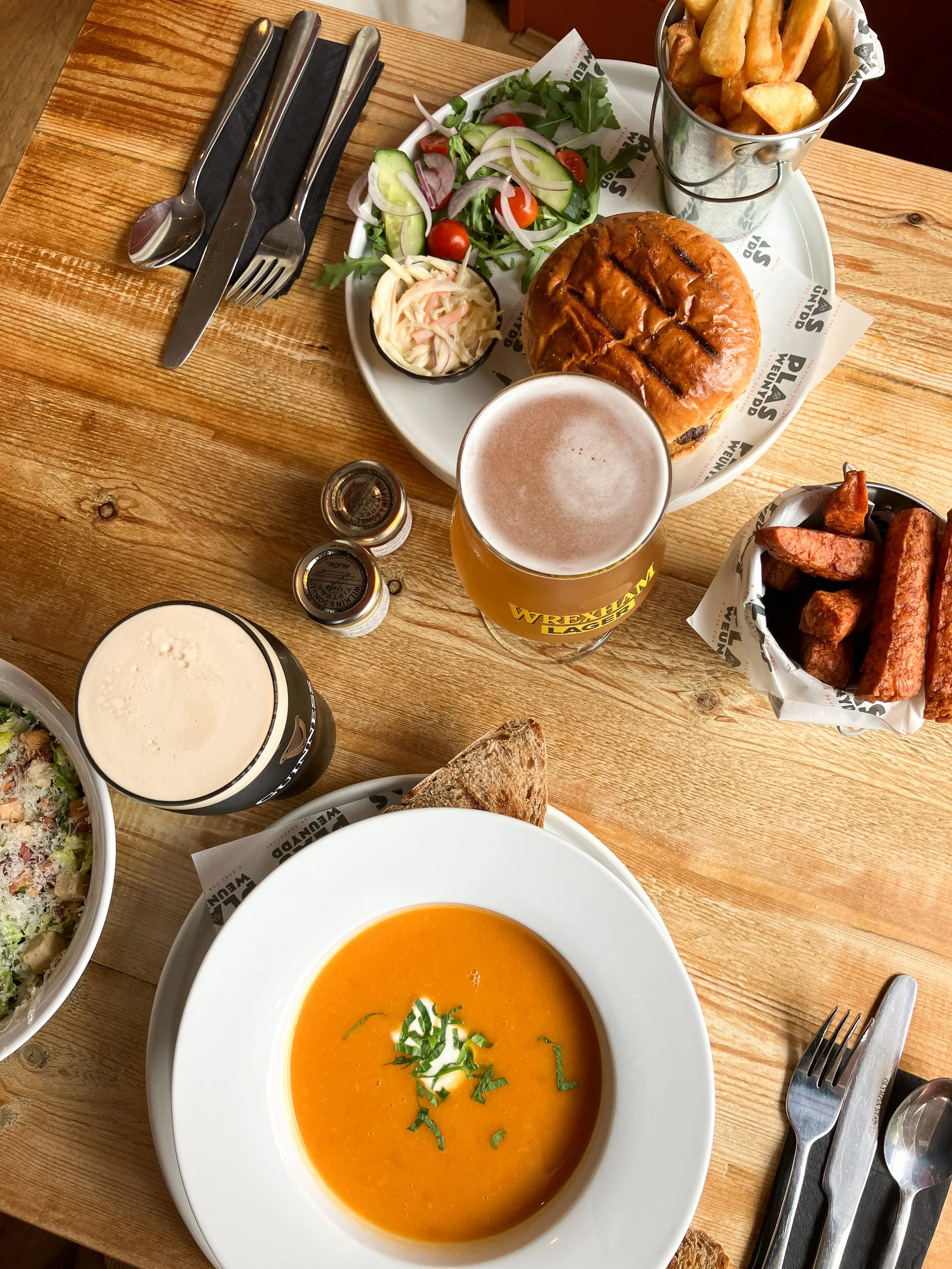 A spread of food on a wooden table, including a bowl of orange soup garnished with herbs, a salad with cherry tomatoes and cucumbers, a burger with grill marks, a glass of beer, a can of beer, a glass of dark beer, and side dishes of fries and sweet potato wedges.