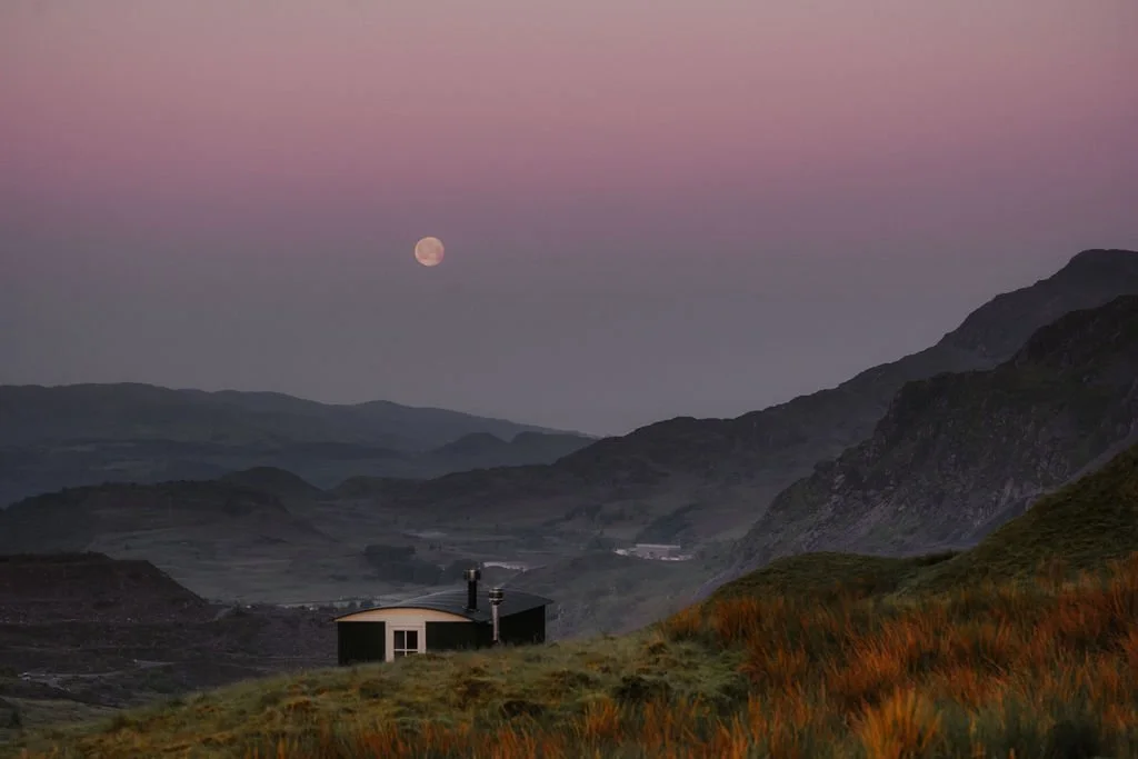 Sunset over rolling hills with a small house in the foreground and mountains in the background, moon visible in the sky.