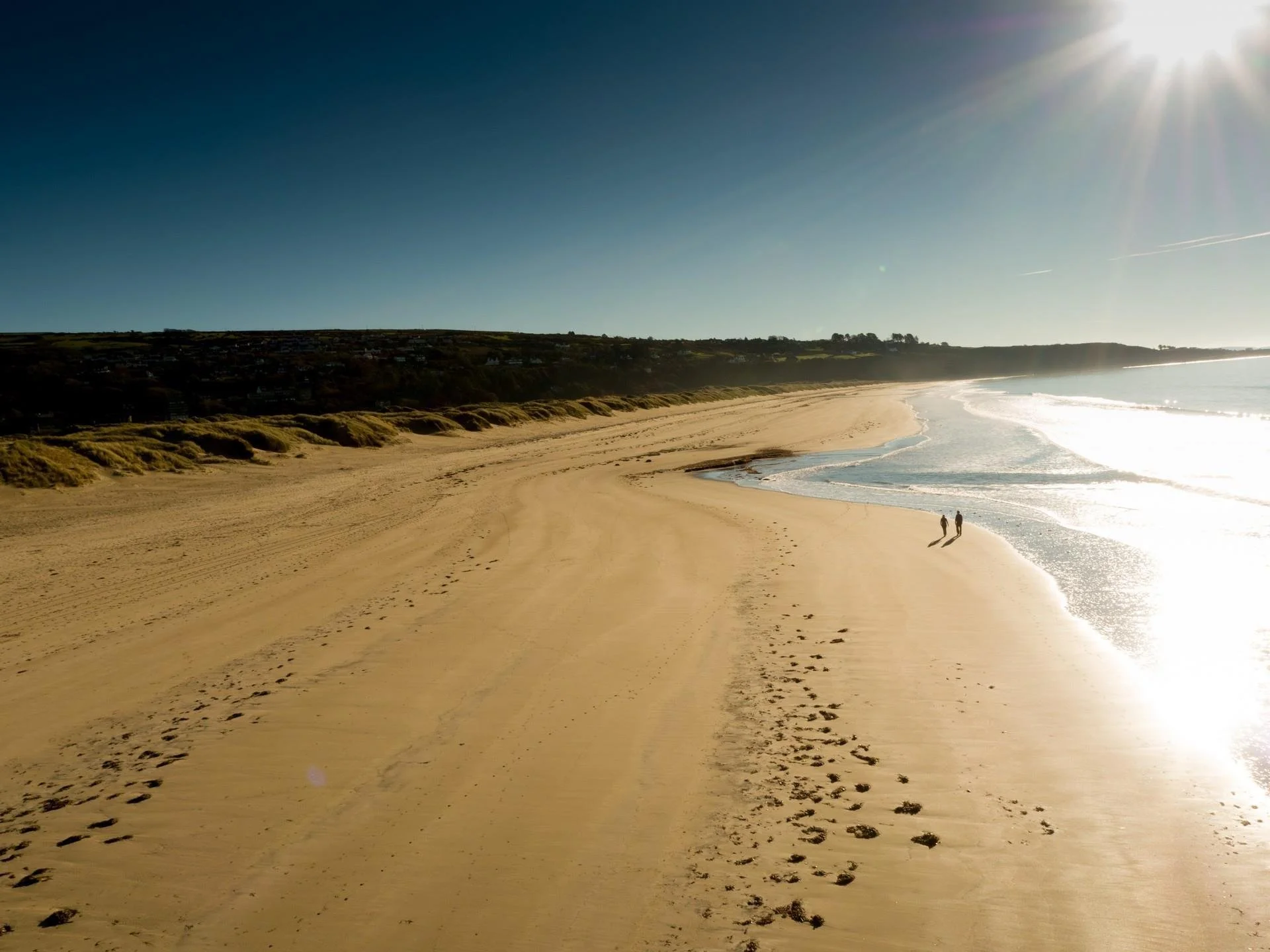 A sandy beach with footprints and two people walking, with cliffs in the background, sunlight shining from the top right corner.