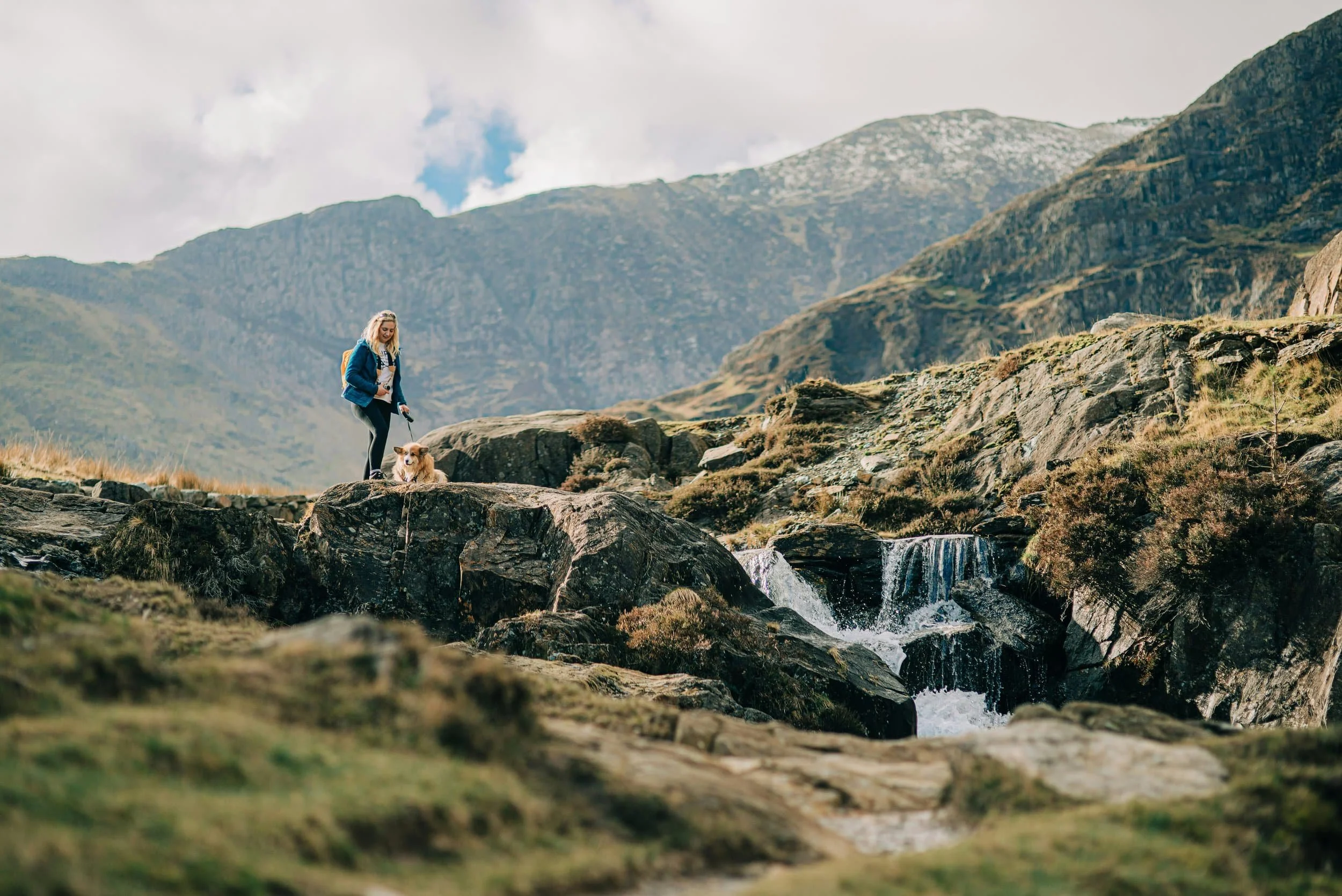 A woman hiking with her dog on a rocky trail near Plas Weunydd in a mountainous area with grassy patches, rocks, a small waterfall, and mountains in the background under a partly cloudy sky.