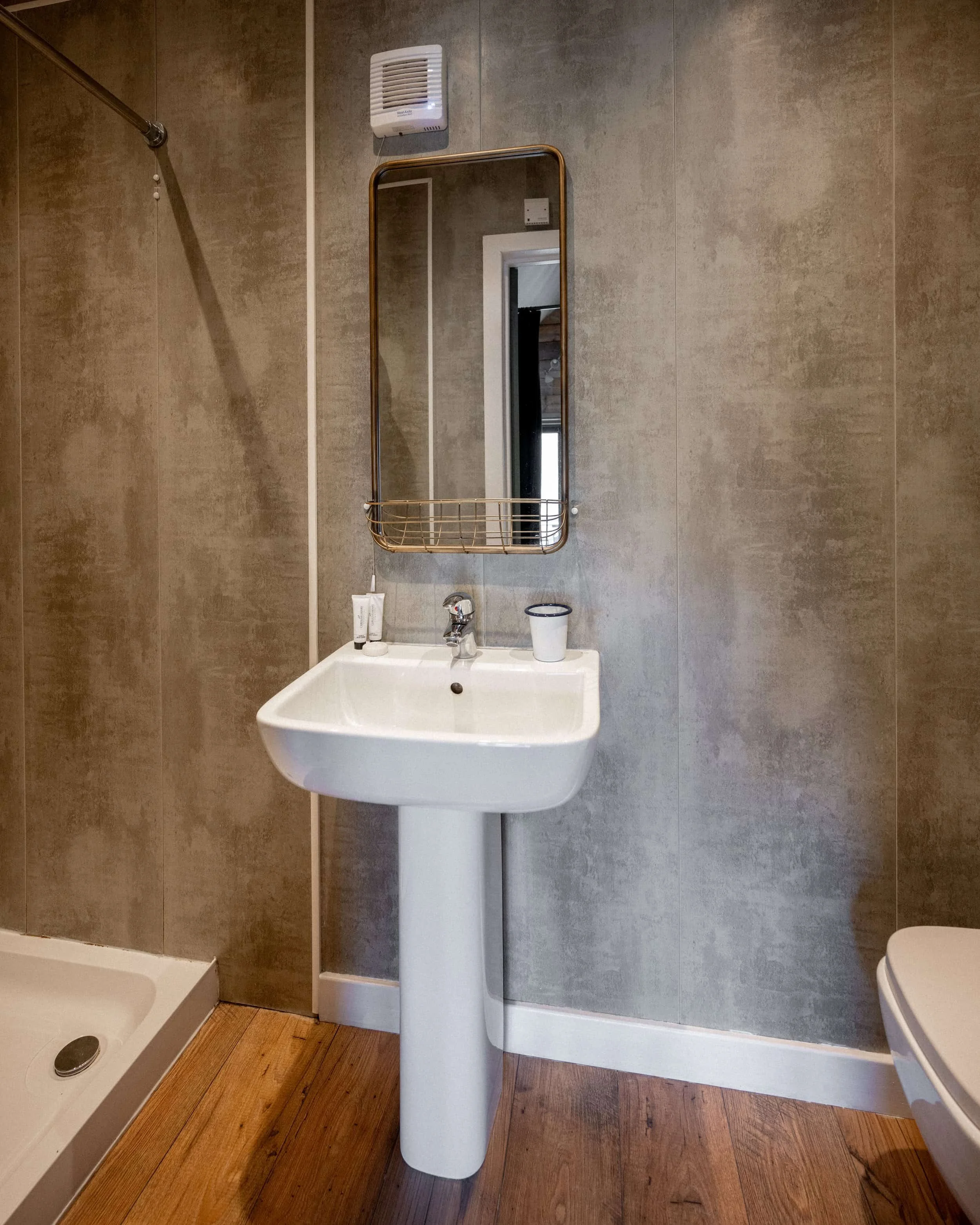 A bathroom inside the glamping pod with a white sink, a mirror above it, a small shelf, and a toilet on the right. The walls are gray, and the floor is wooden. There is a vent near the ceiling and a shower area to the left.