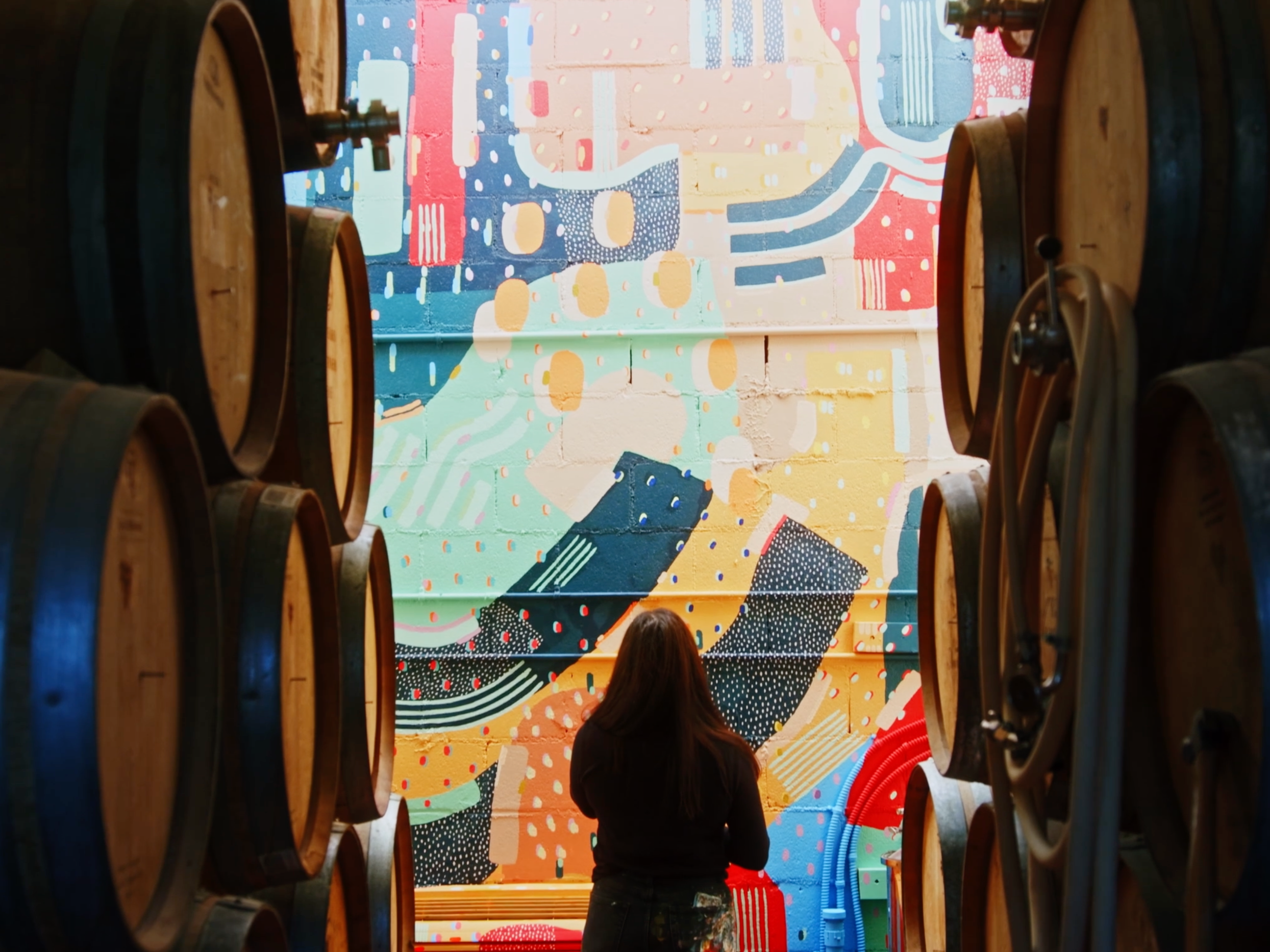 Beau Kester Cinematographer  Julu stands facing a colorful abstract mural, framed by wine barrels in a wine cellar.