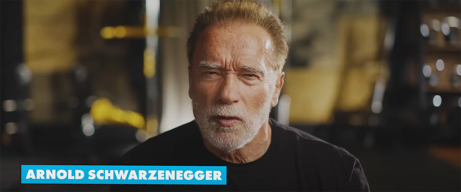 Beau Kester Director and Director of Photography capturing still from The Britto Doc documentary about Romero Britto’s, Arnold Schwarzenegger with a serious expression, wearing a black shirt, in a gym setting with blurred equipment in the background.