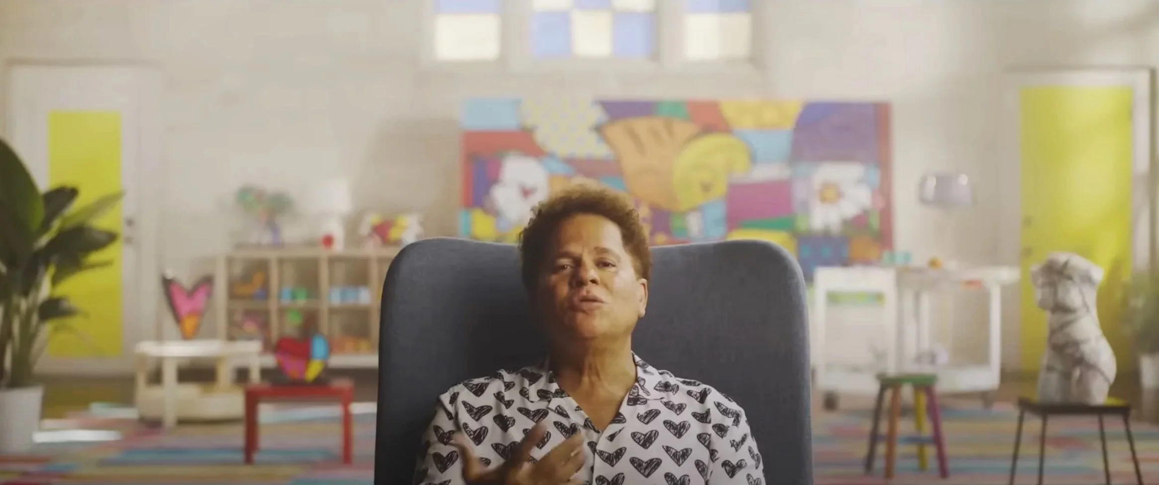 Beau Kester Director and Director of Photography capturing still from The Britto Doc documentary about Romero Britto. Romeros sits for first interview in colorful room with artwork, plants, and sculptures, speaking and gesturing with her right hand.