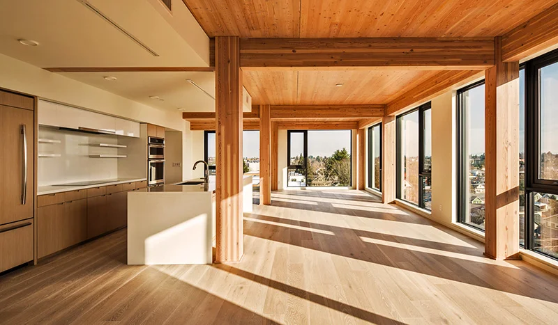 Above: The combination of exposed CLT wooden ceilings and exposed glulam posts and beams on a grid with oak floors lends a warm wooden patina to the units, offset by vertical white drywall panels between the windows on the three sides of the unit an…