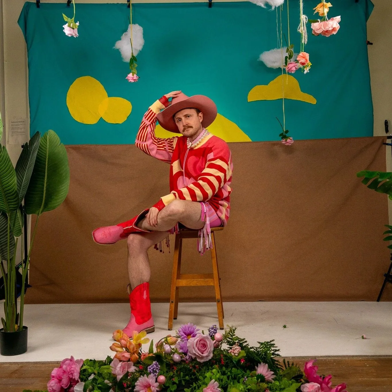 Man dressed in colorful, floral eccentric costume sitting on a stool surrounded by flowers, plants, and a cheerful backdrop depicting a sky with clouds and bright yellow shapes.