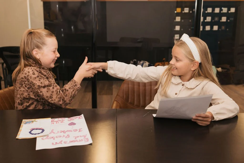 Two young girls sitting at a table, smiling and playfully touching fingers, with one girl holding a handwritten note, in an indoor setting
