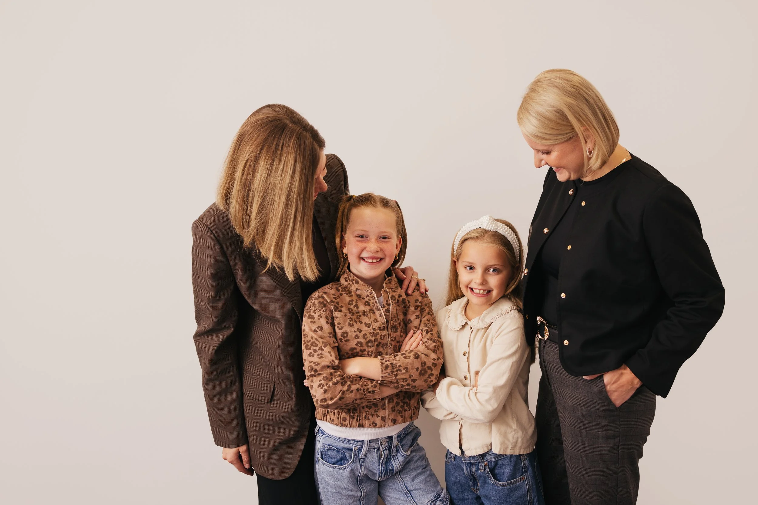 Two adult women and two young girls smiling and looking at each other against a plain wall.