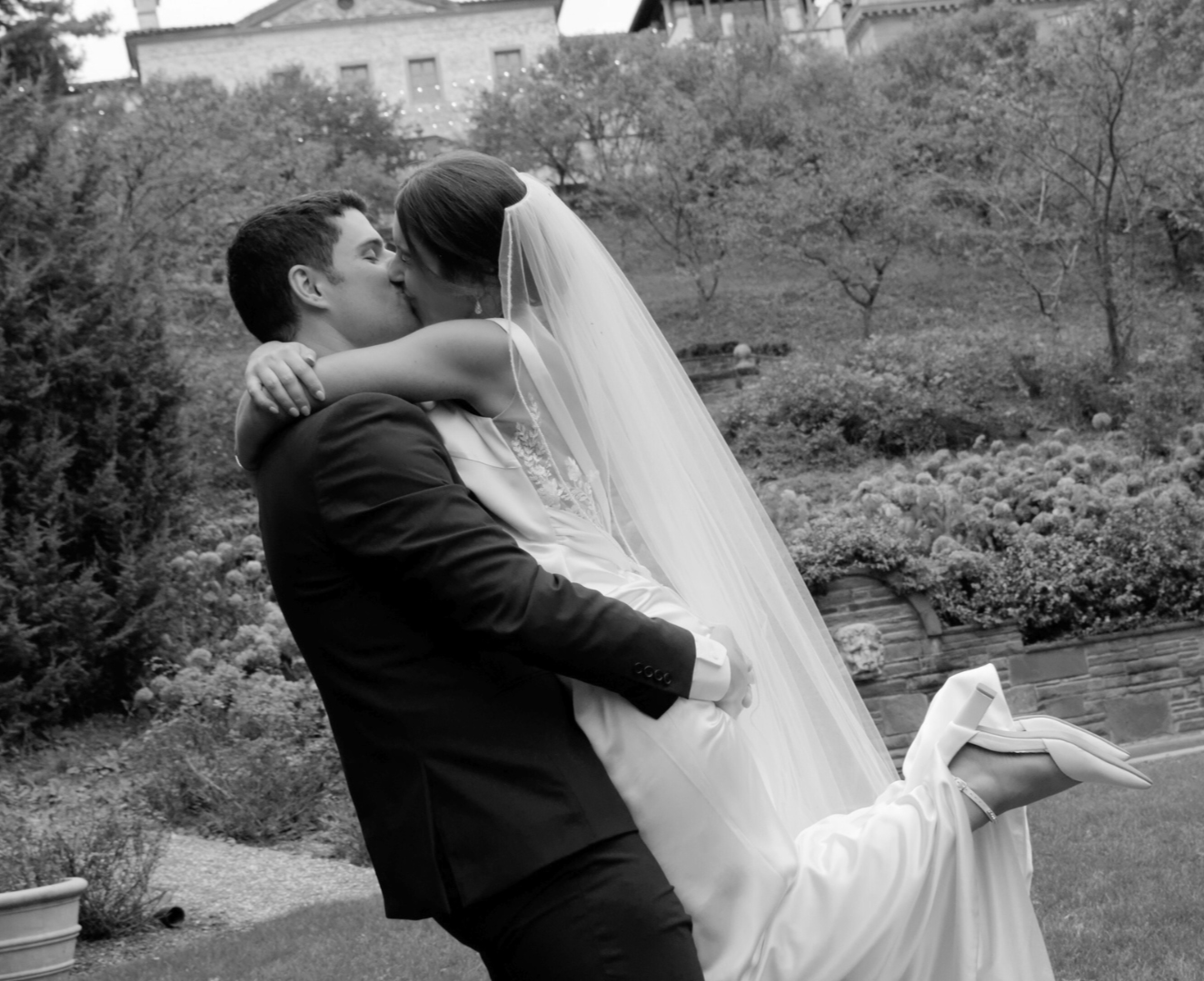 A groom holding a bride, who is wearing a wedding dress and veil, in an outdoor garden setting.