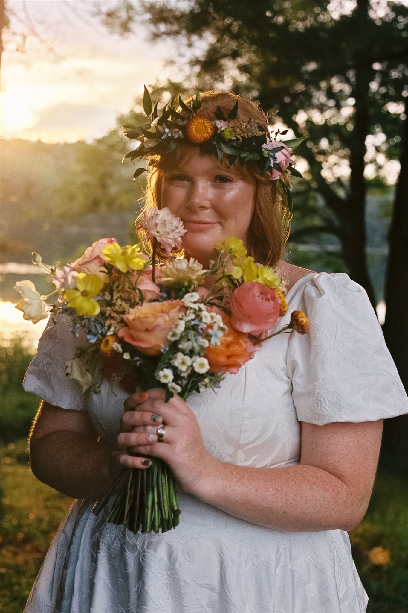 Woman in a white dress holding a bouquet of flowers, wearing a flower crown, outdoors during sunset.