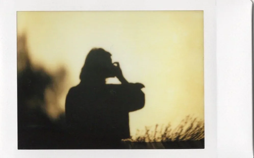 Silhouette of a person wearing glasses and a hat, sitting outdoors with a hand on their face, against a yellow sky at sunset or sunrise.