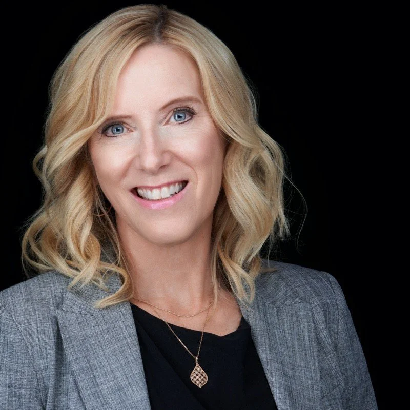 A professional woman with blonde, wavy hair, blue eyes, wearing a gray blazer over a black top, with a gold pendant necklace, smiling against a black background.