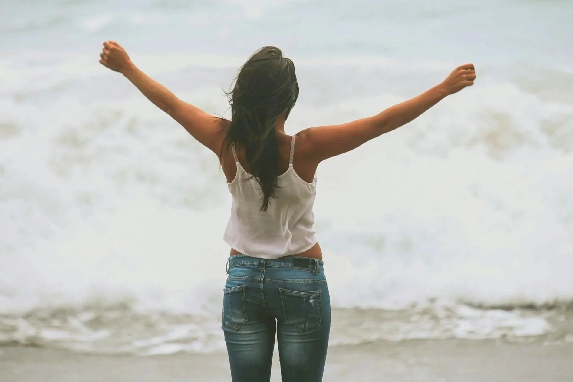 A woman with long dark hair wearing a white tank top and jeans standing on a beach with arms raised, facing the ocean waves.