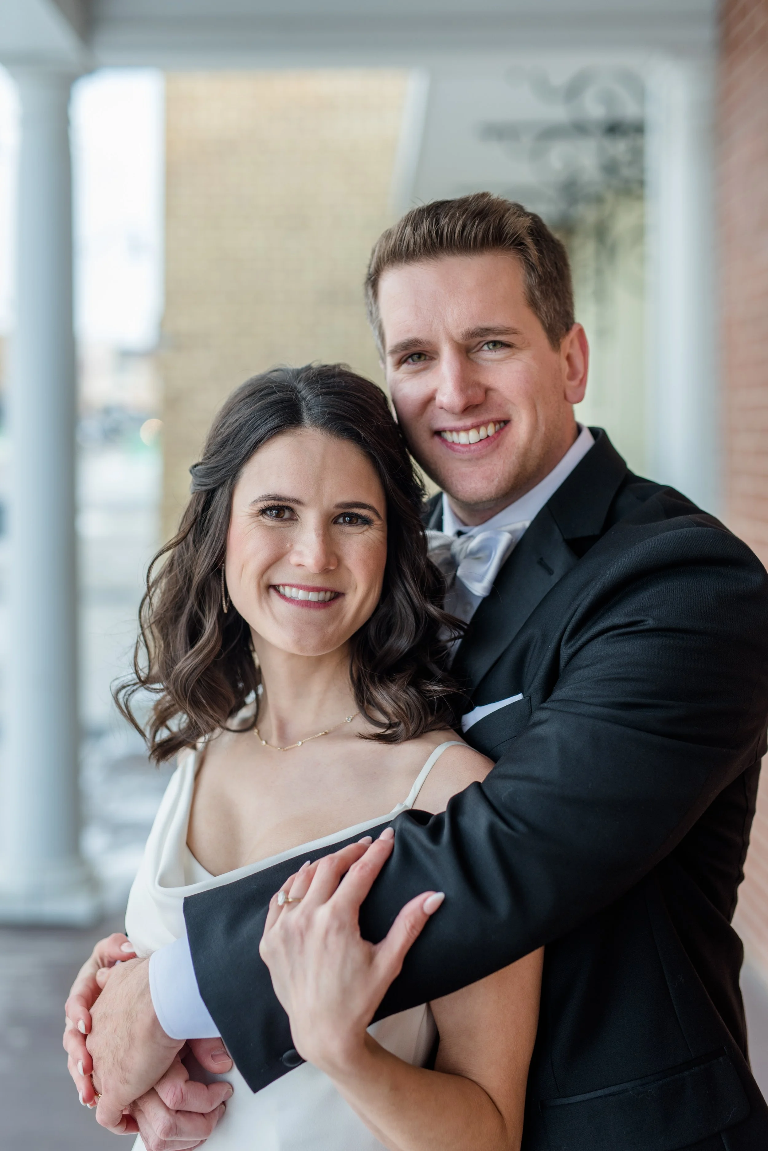 Groom hugs bride from behind as they pose on the covered porch at the Broz in New Prague, Minnesota