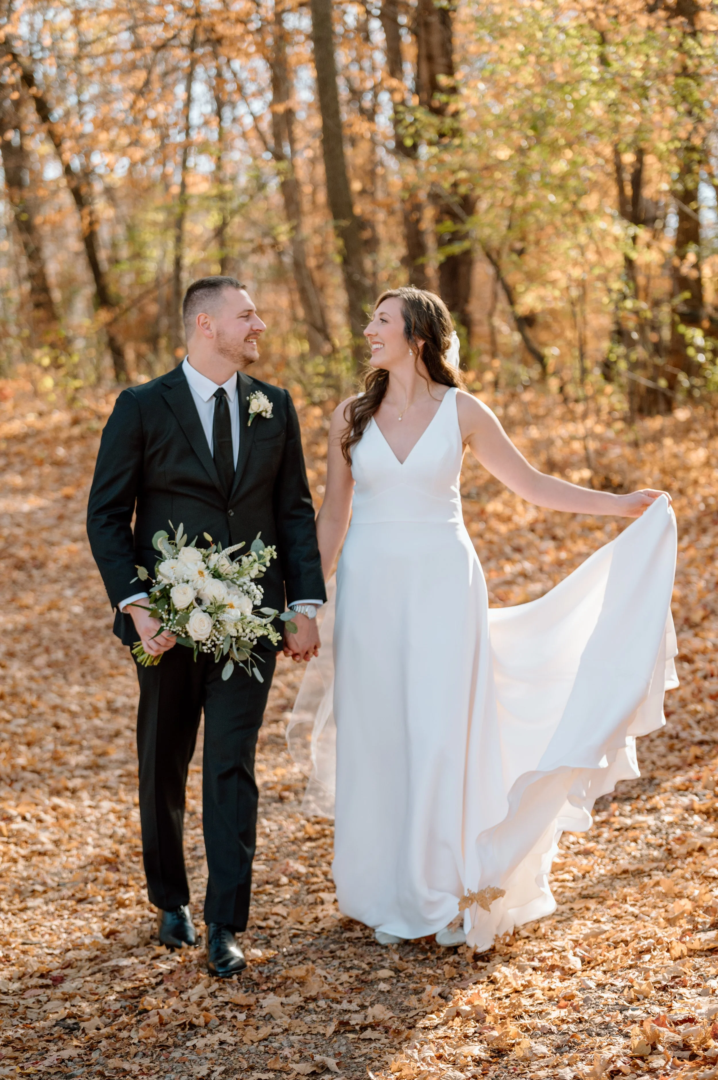 Bride and groom hold hands and walk through the forest as golden leaves surround them on the trees and on the ground on their fall Minnesota wedding day