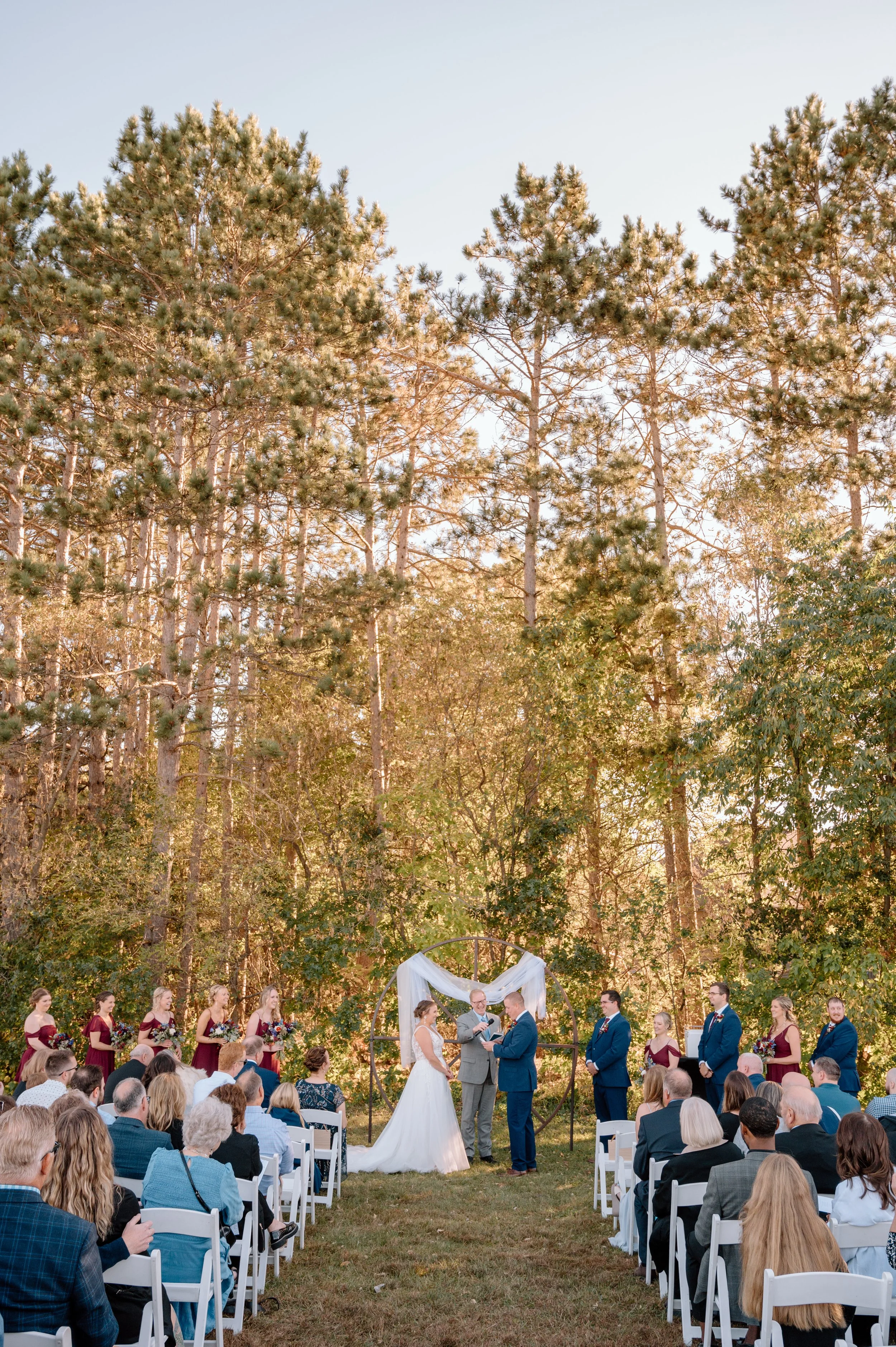 Bride and groom exchange vows in front of wagon wheel draped with white fabric in a ceremony in front of towering pine trees at Stone Lion Winery