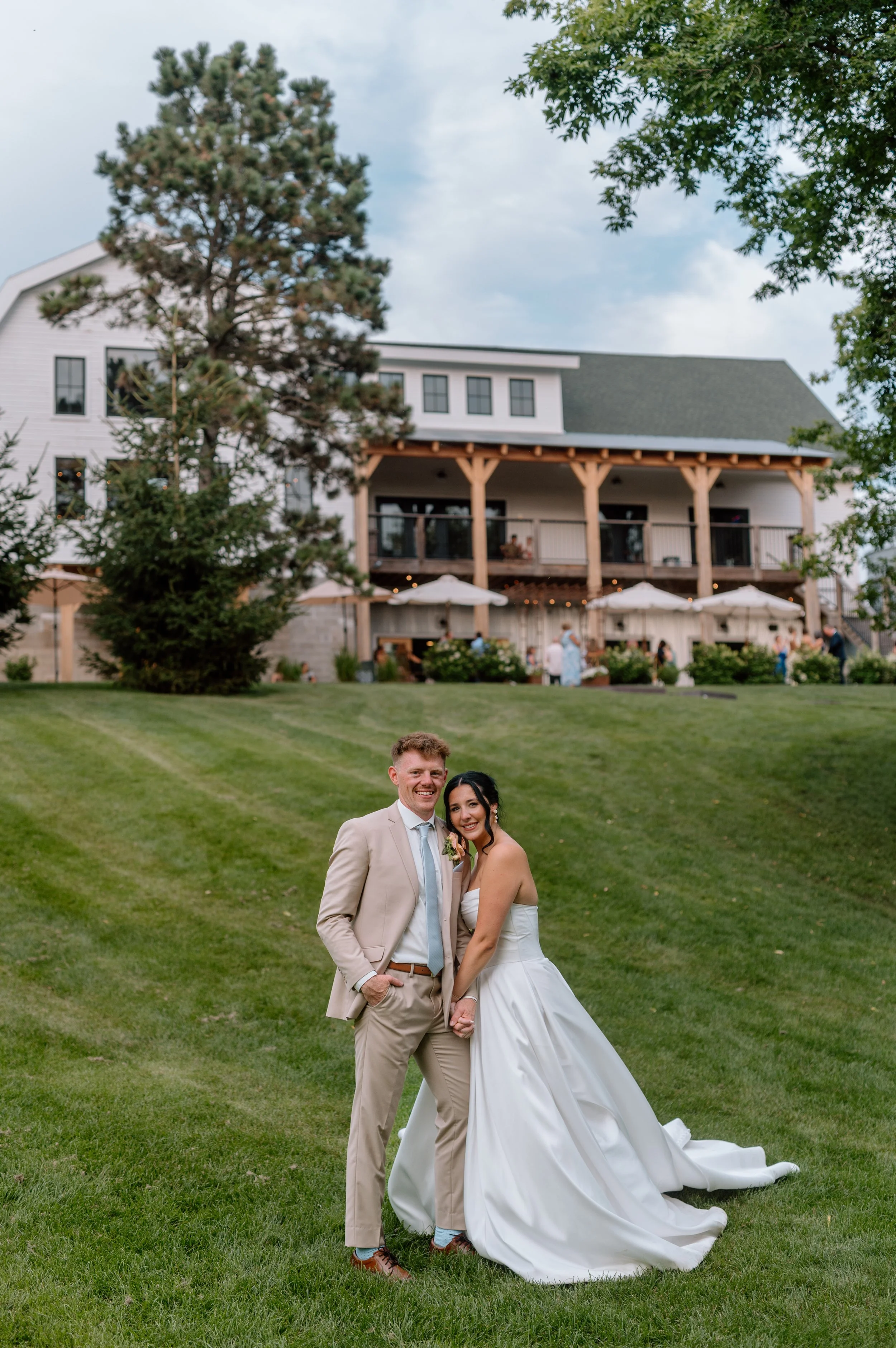 Alexandria, MN wedding venue Gathered Oaks during cocktail hour as bride and groom hold hands and smile at the camera on the back lawn with the white barn behind them