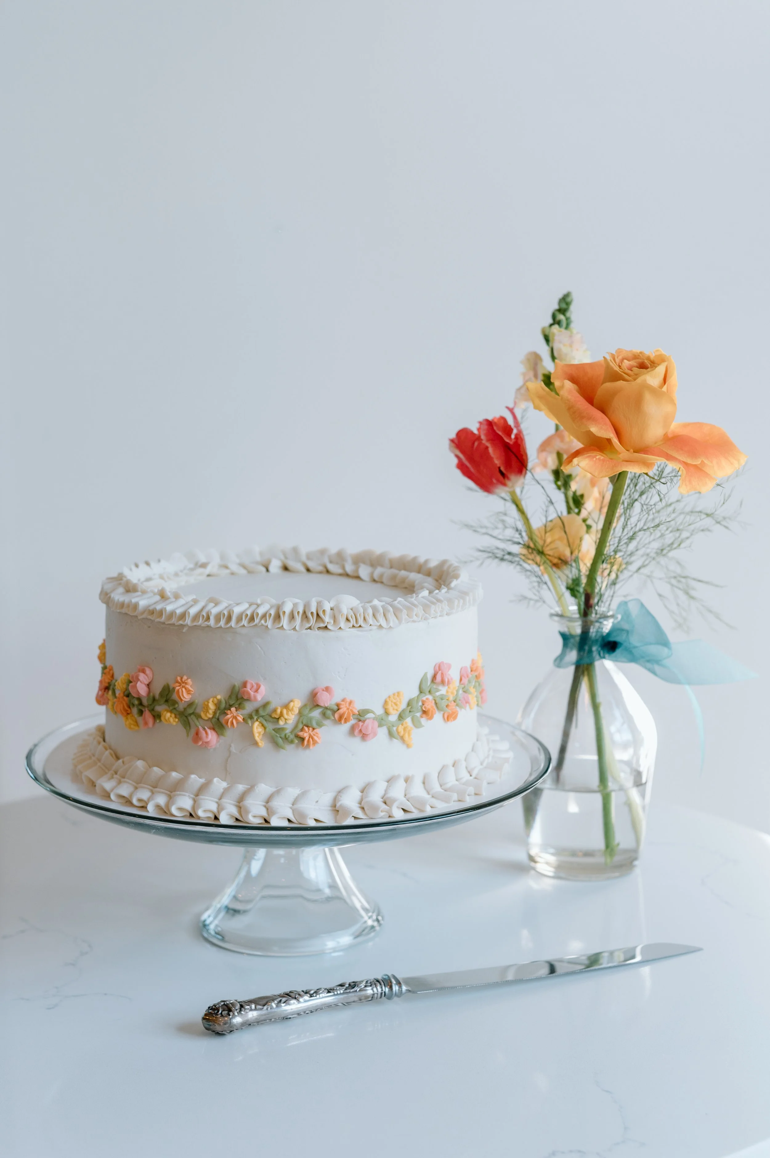Simple and classic single tiered wedding cake with sunset colored flowers piped around the outside sits on a glass cake stand in front of sunset colored flowers at summer Belle Ame wedding day