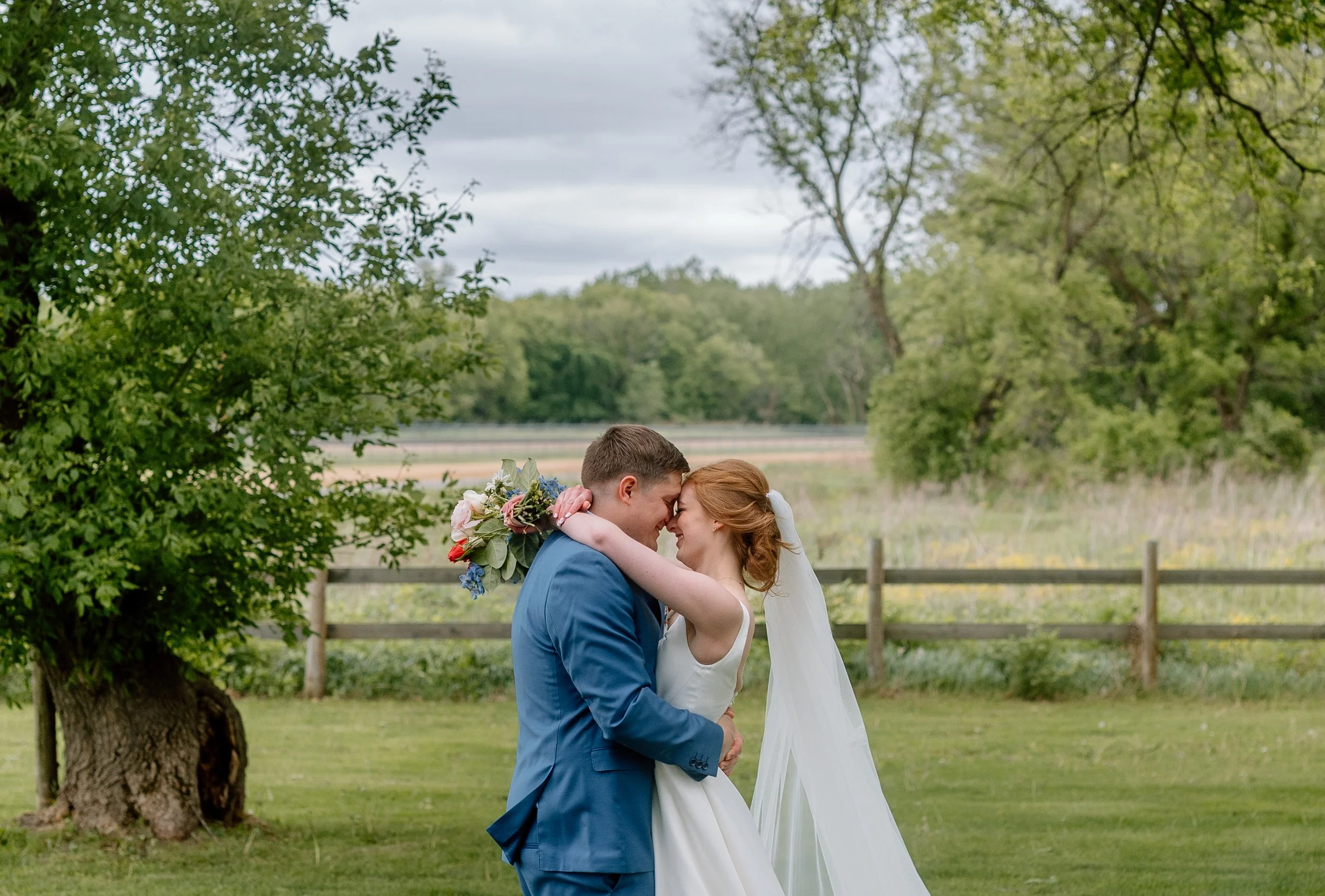 Bride and groom embrace, foreheads together, with trees, an open field, and rustic wooden fence behind them on their wedding day at Genesis Ranch in Rosemount, MN
