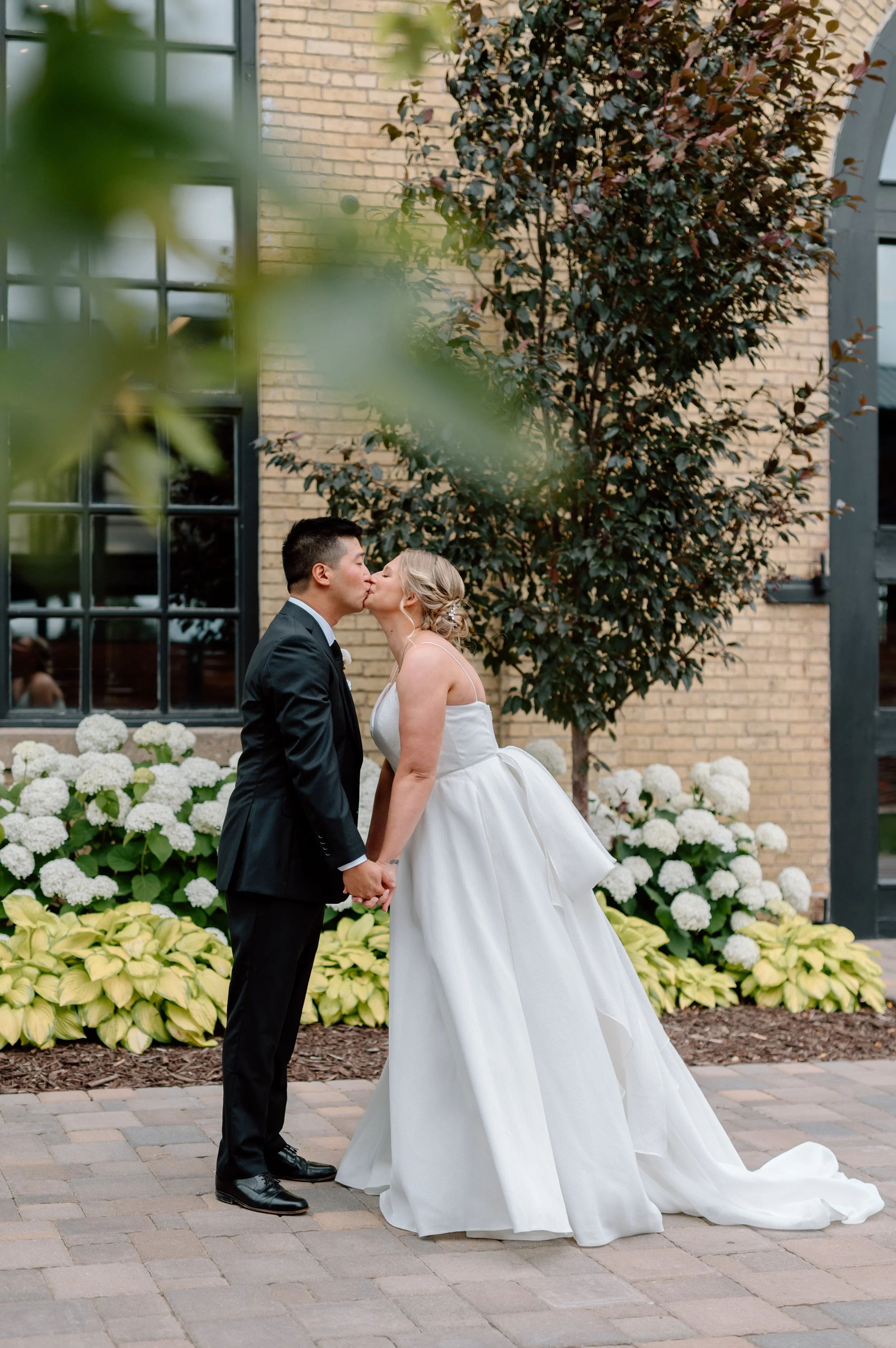 Bride and groom share a kiss during their first look in front of white hydrangeas outside St. Paul wedding venue The Essence Event Center