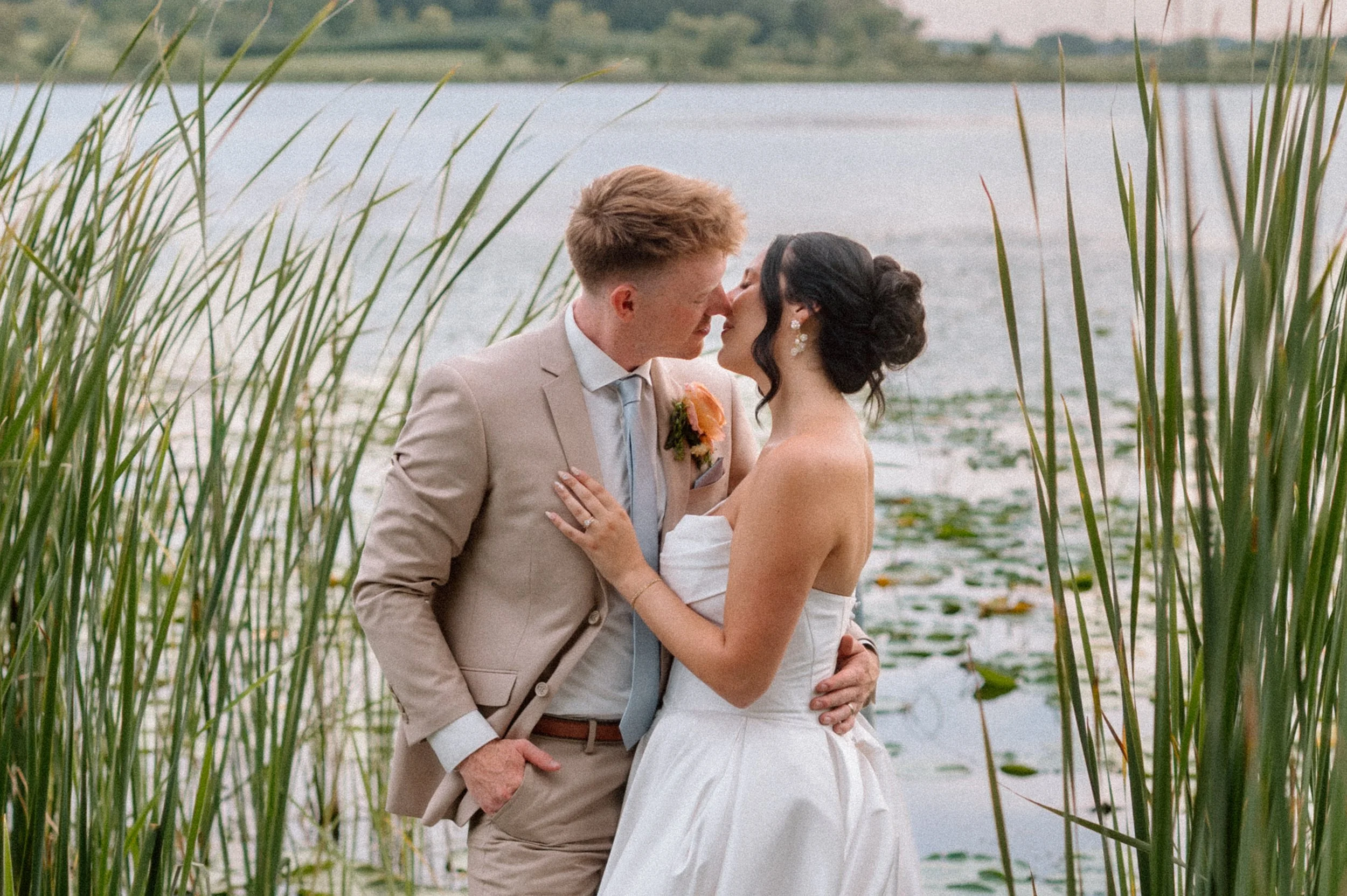 Bride and groom kiss in front of the pond at sunset on their summer wedding day at Gathered Oaks in Alexandria, MN