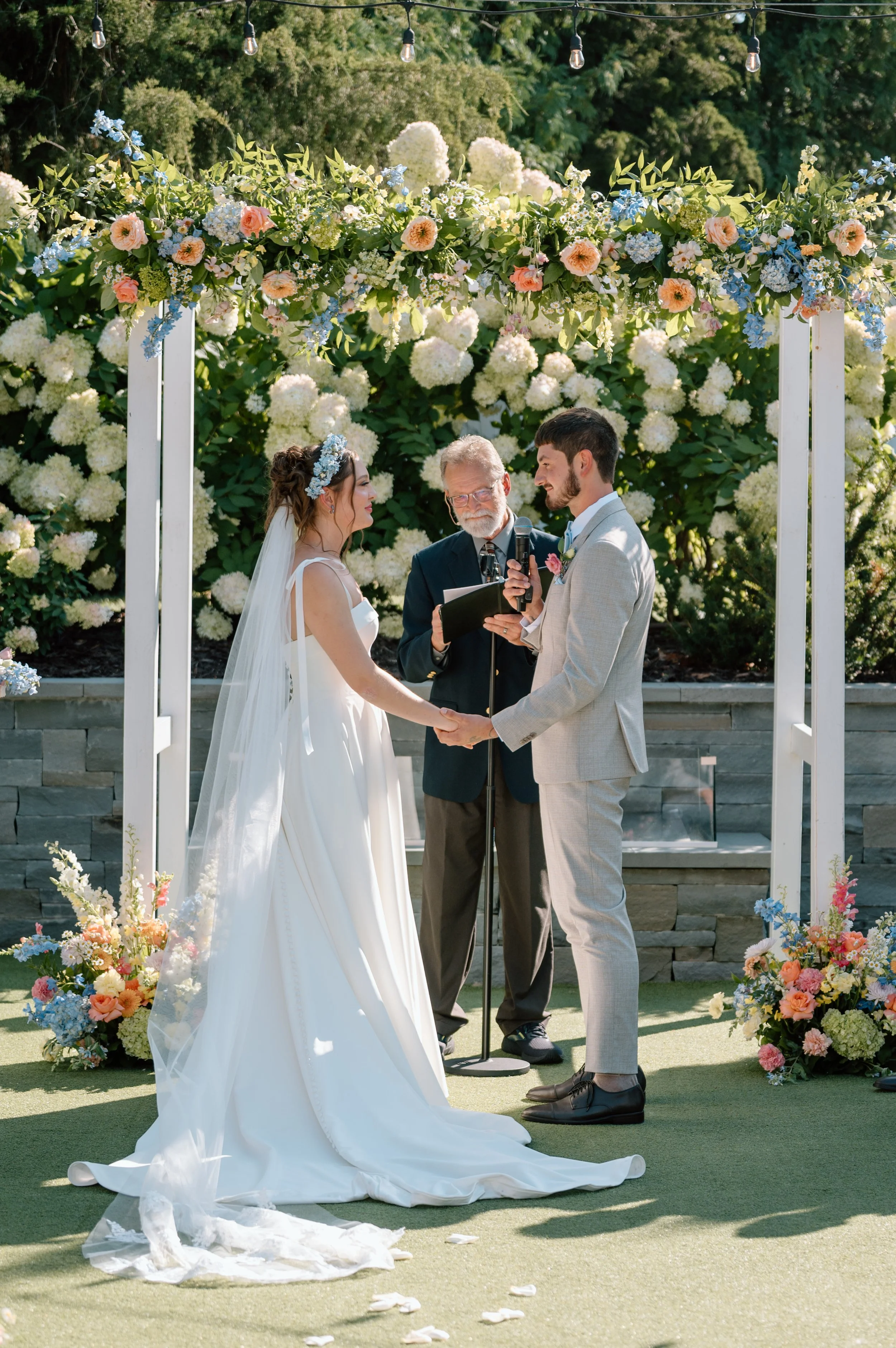 Bride and groom exchange vows under a white arch decorated with colorful florals in front of white hydrangeas in the courtyard of the Hutton House, MN
