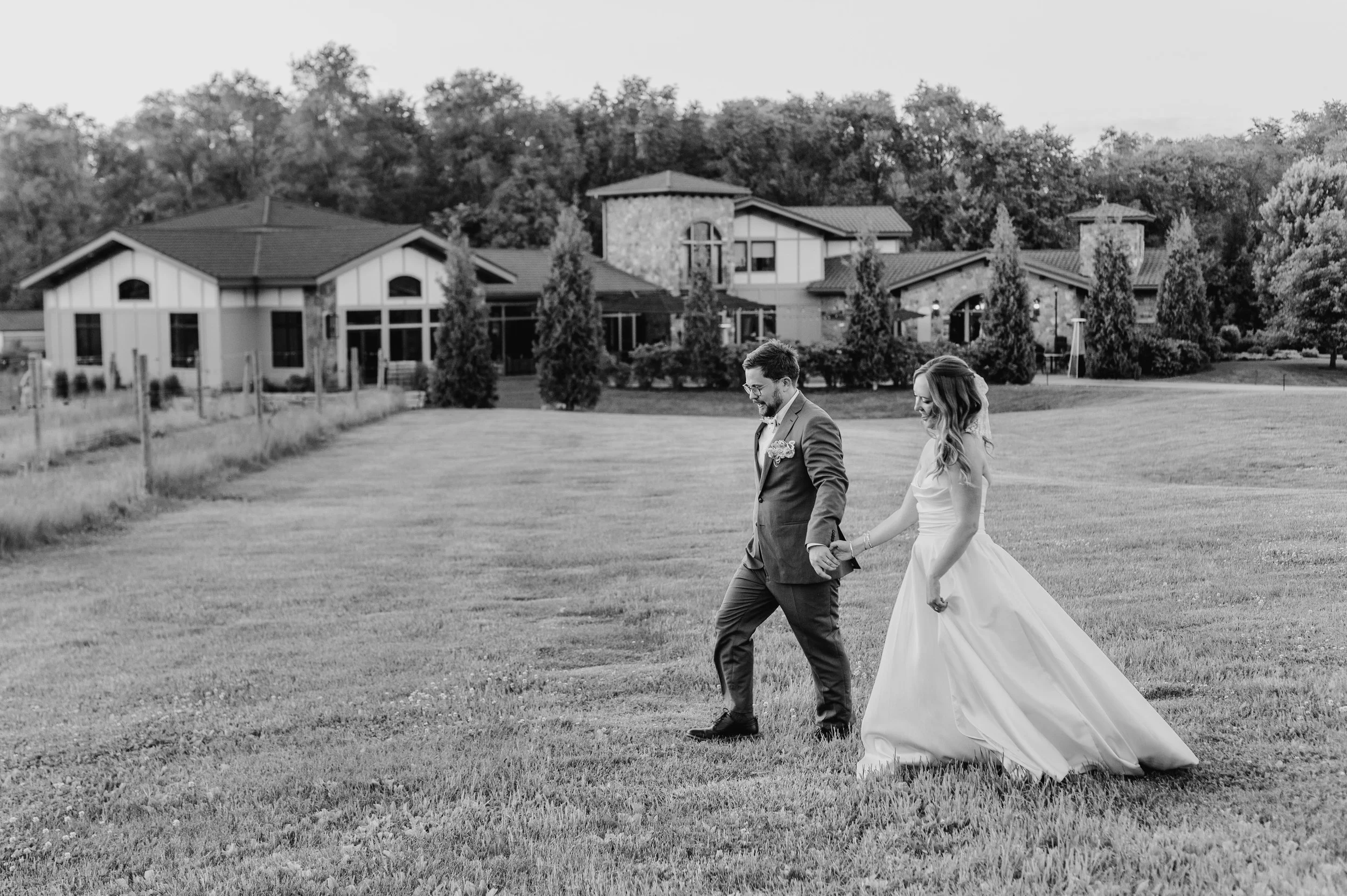 Black and white image of groom holding bride's hand and guiding her on a walk across the grass beyond Belle Ame wedding venue near the Twin Cities; MN wedding photographer