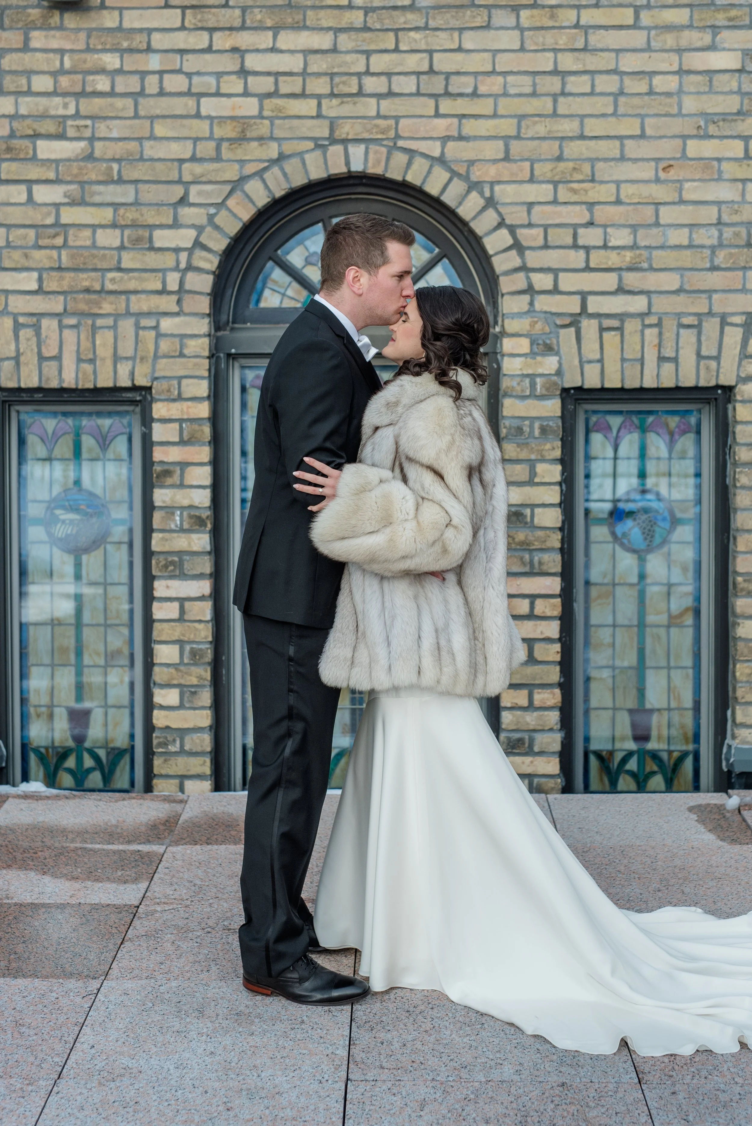 Groom kisses's bride's forehead in front of stained glass window on the secret patio at The Broz in New Prague, Minnesota