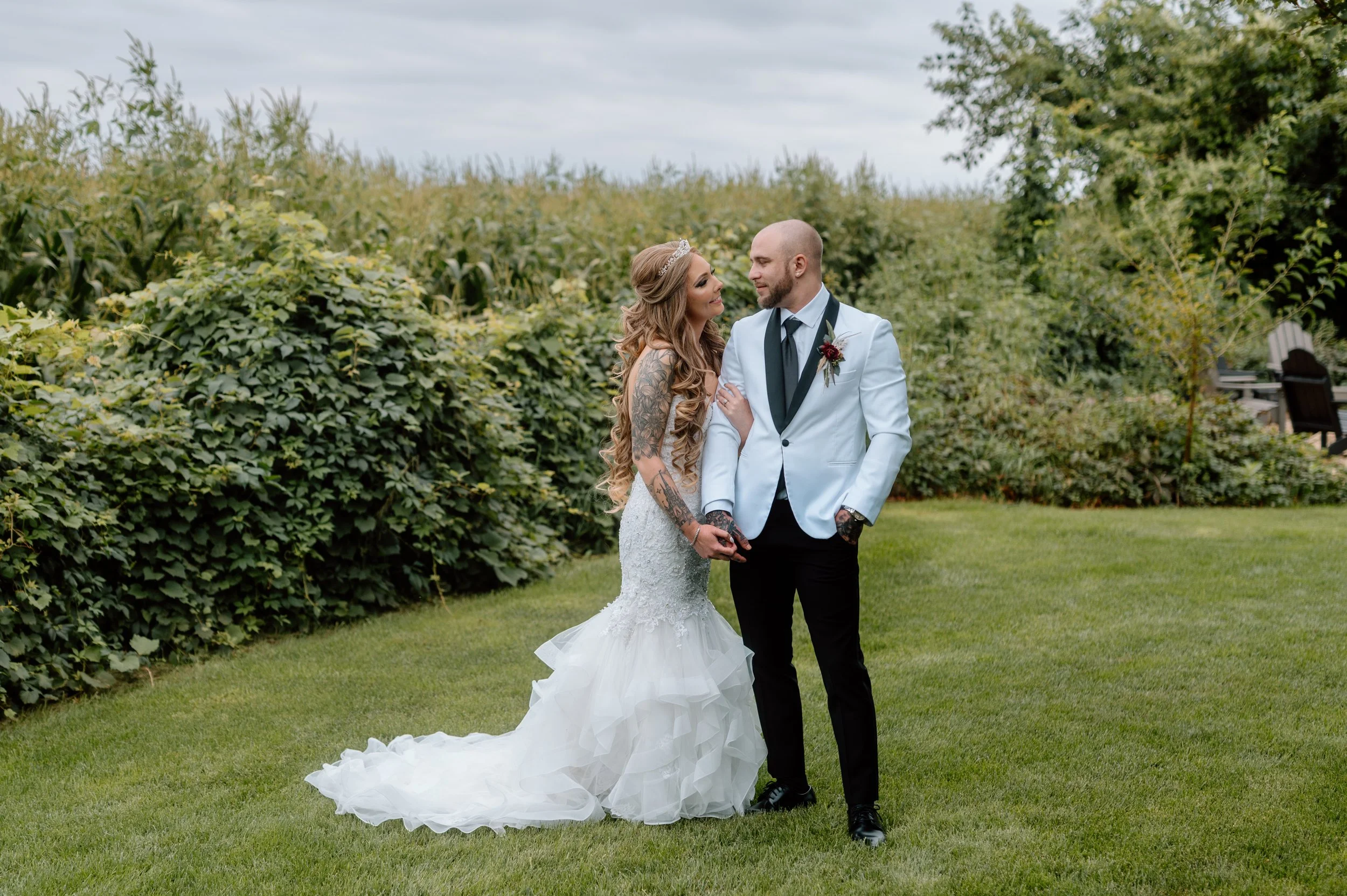 Tattooed bride and groom link arms and gaze at each other in front of greenery and vines at Minnesota wedding venue Legacy Hill Farm