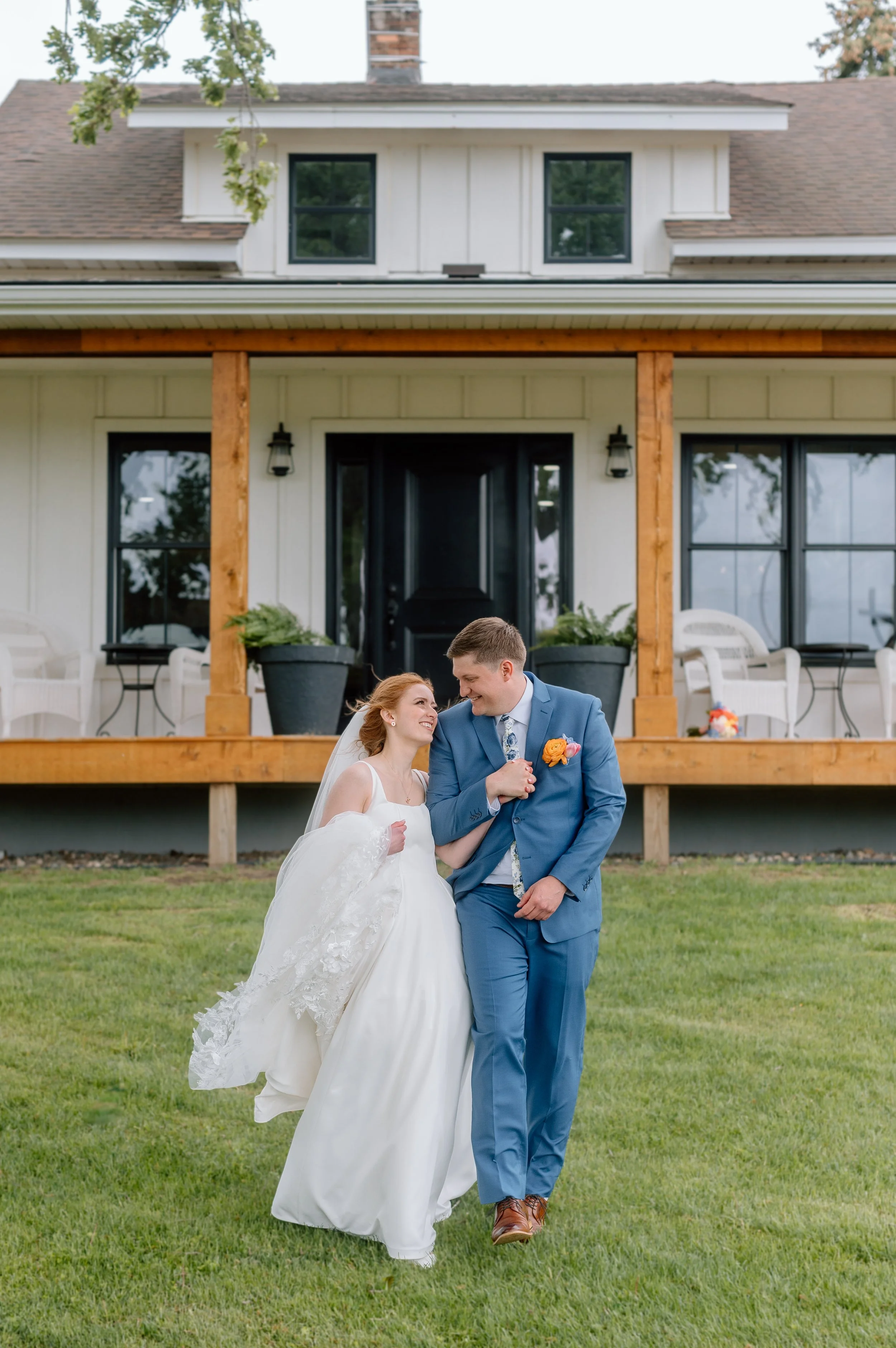 Bride and groom hold hands, look at each other, and walk across the lawn in front of the house at Genesis Ranch, MN