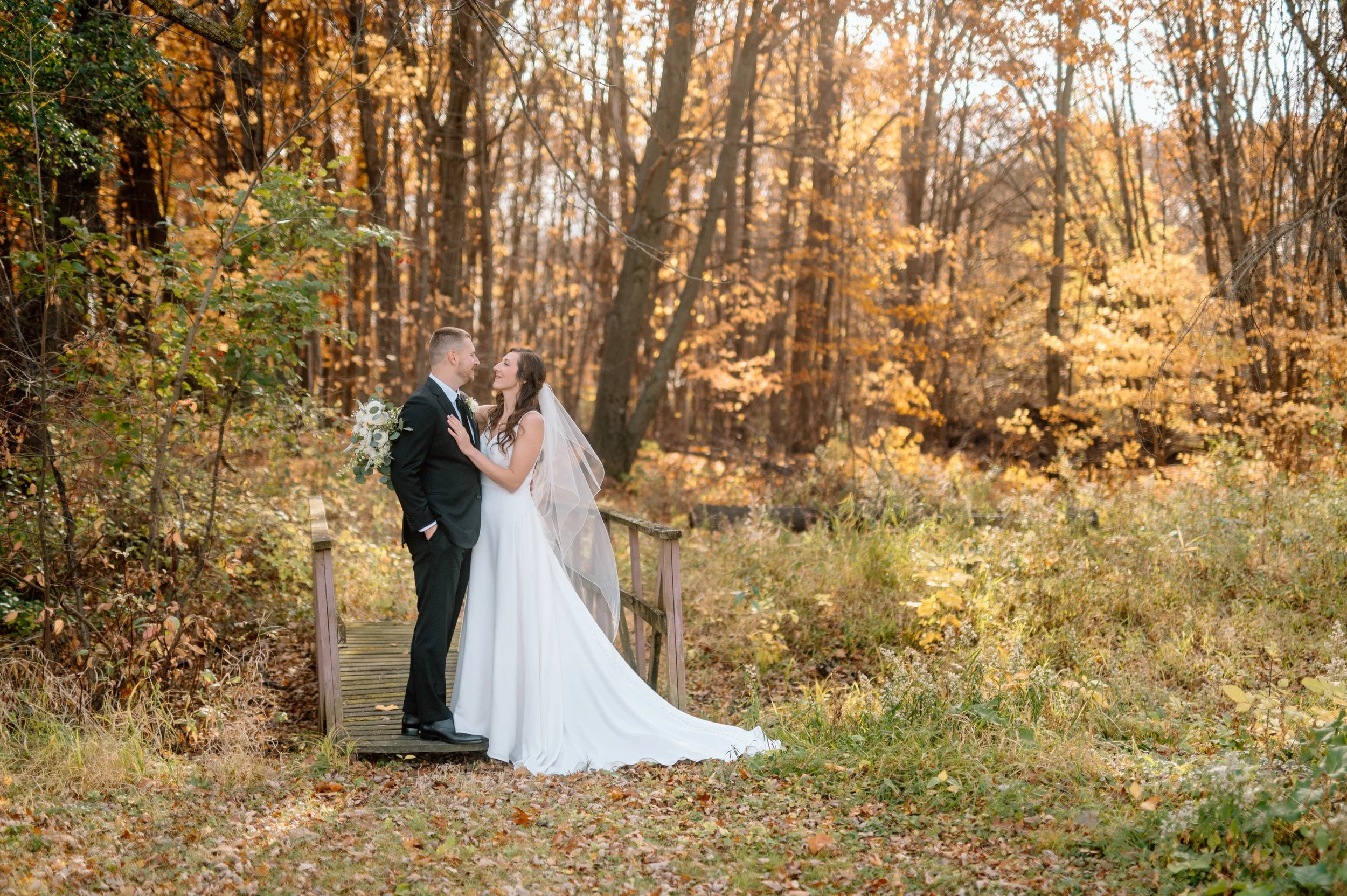Bride and groom stand on small wooden bridge while looking at each other, surrounded by golden fall leaves on Minnesota wedding day in the Twin Cities