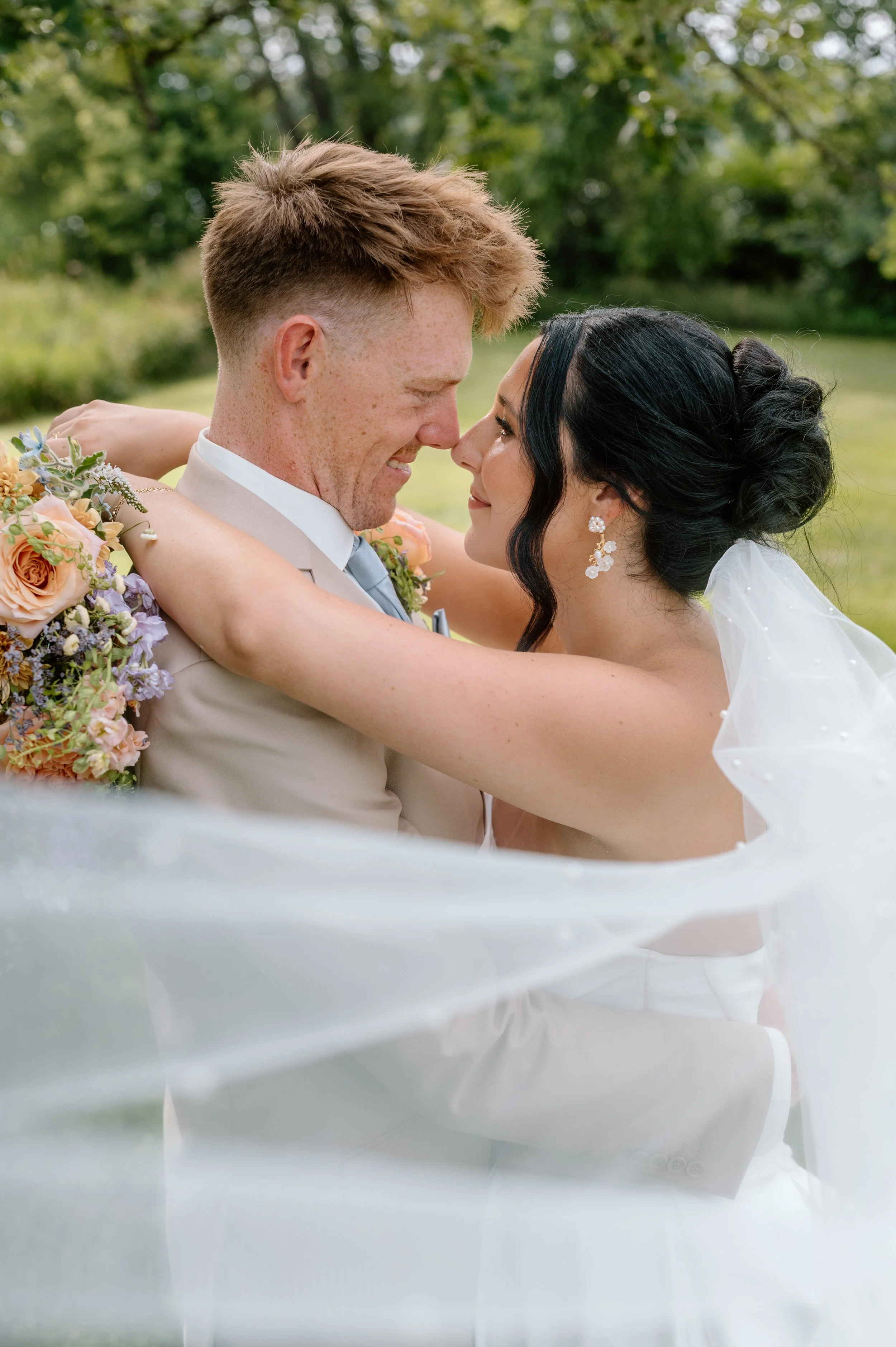 Bride and groom stand nose to nose and smile at each other from within bride's cathedral-length veil on their wedding day at Gathered Oaks in Alexandria, Minnesota
