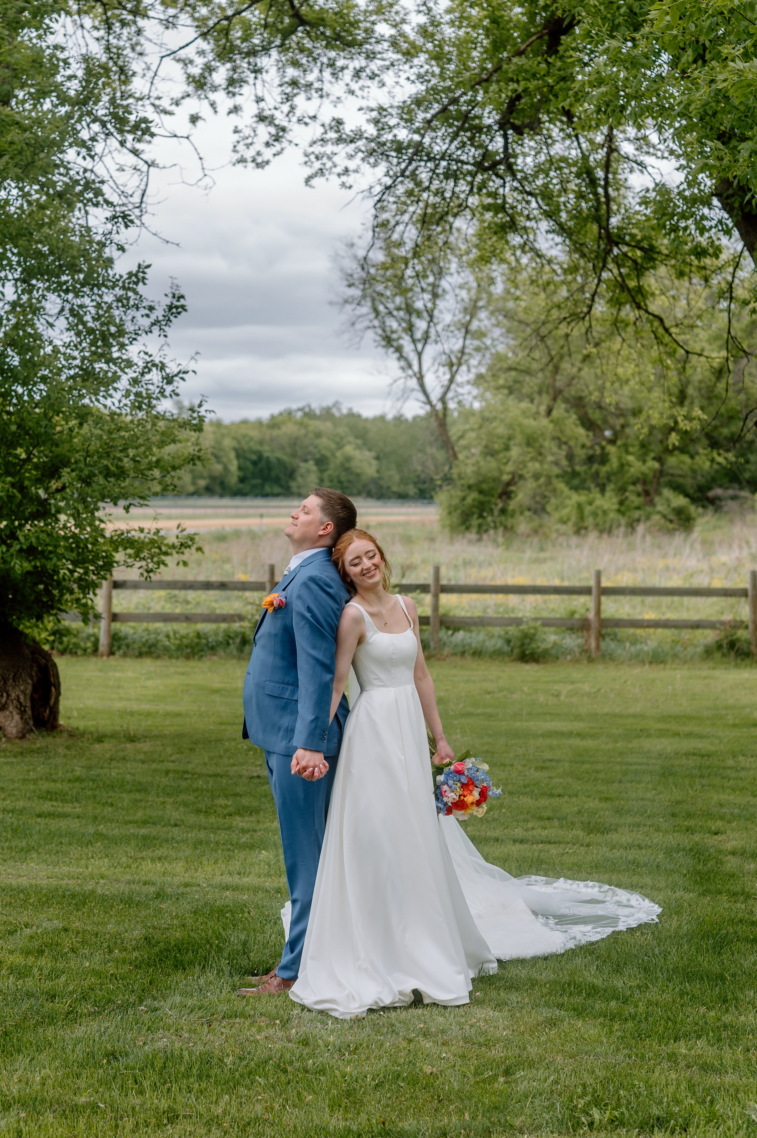 Bride and groom stand back to back in an open field with rustic fence in the distance as they take a breath and make the most of their Minnesota wedding day at Genesis Ranch