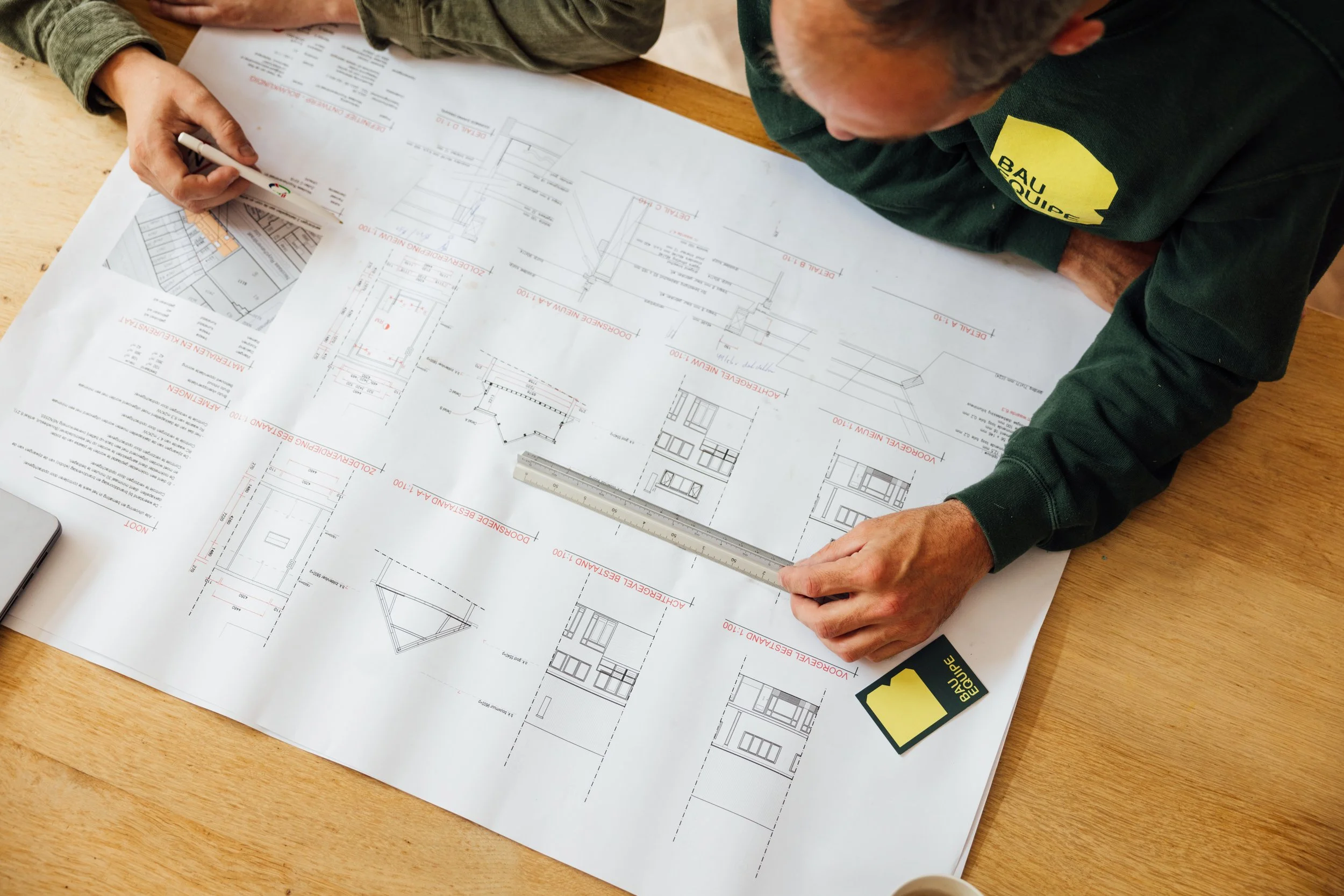 Persoon die bouwtekeningen bestudeert, met een meetlint en een bloemenkaartje met het logo 'BAU Equipe' op een houten tafel. Bau equipe organiseert ook het ontwerpproces, de bouwtekeningen en constructieberekeningen