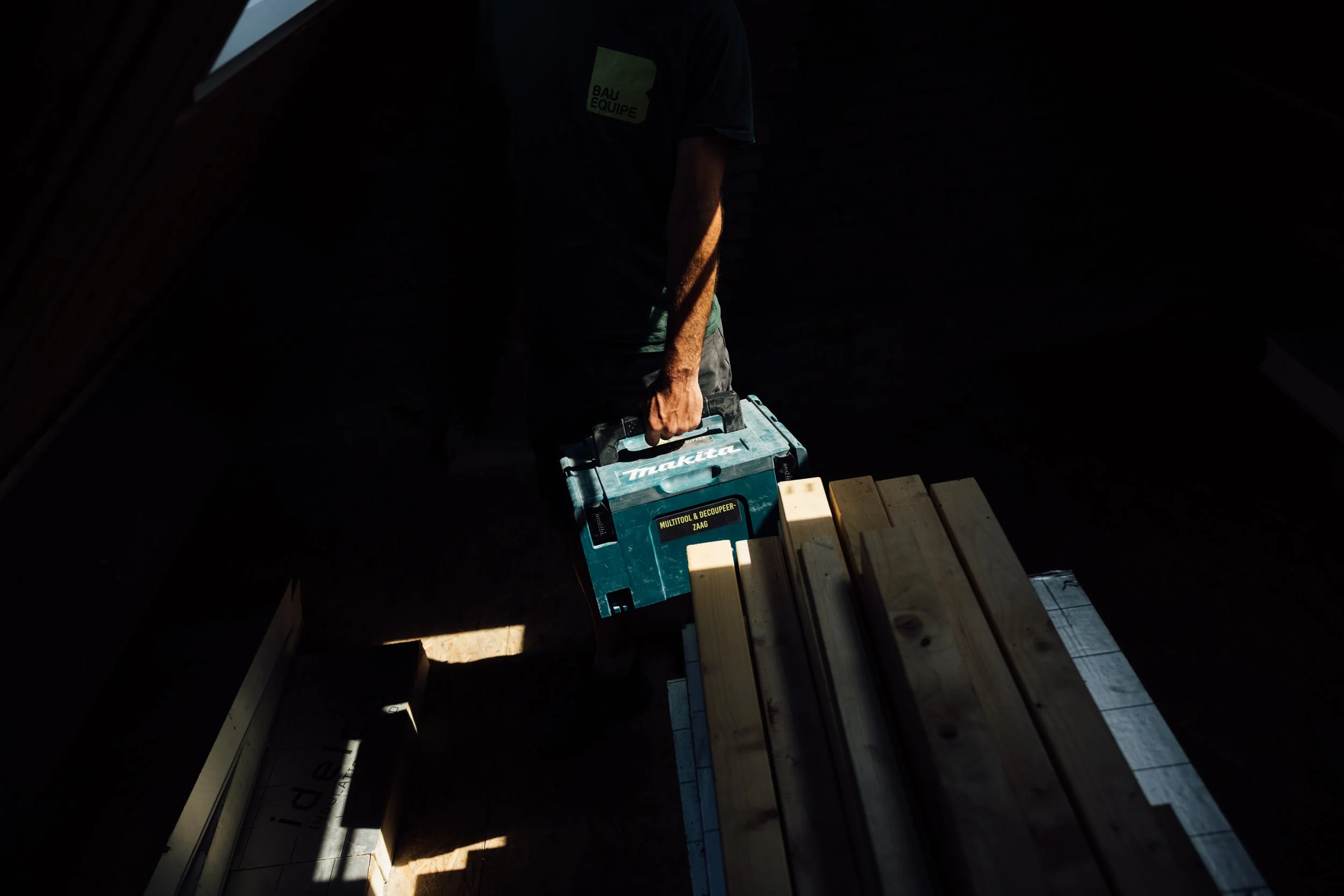 Een persoon die een Makita multitools en decoupeerzaag vasthoudt, in een donkere kamer met sunlight en schaduwen. Holzplanken liggen op een tafel.