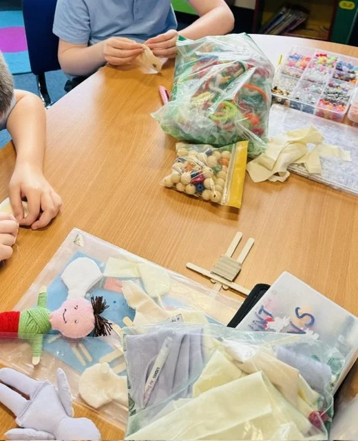Children working on a craft project with various craft supplies including fabric, stuffed toys, beads, and small wooden sticks on a wooden table.