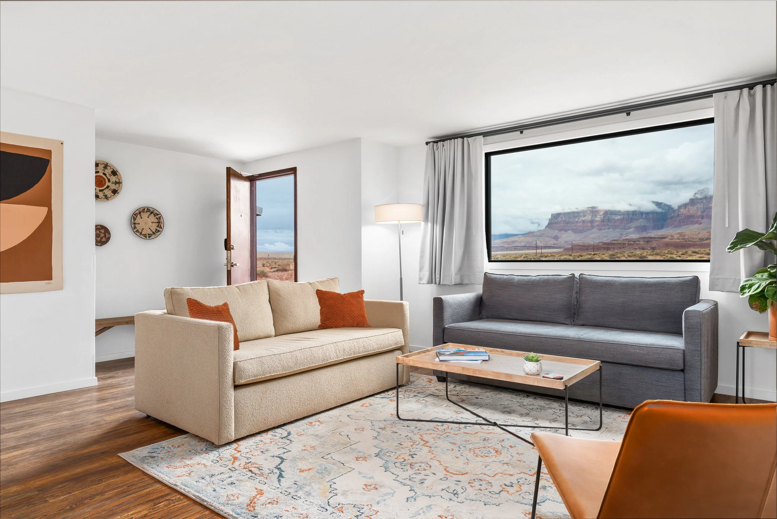 Interior of a living room with two sofas, a coffee table, a floor lamp, a large window showing a desert landscape with mountains, and some wall art.