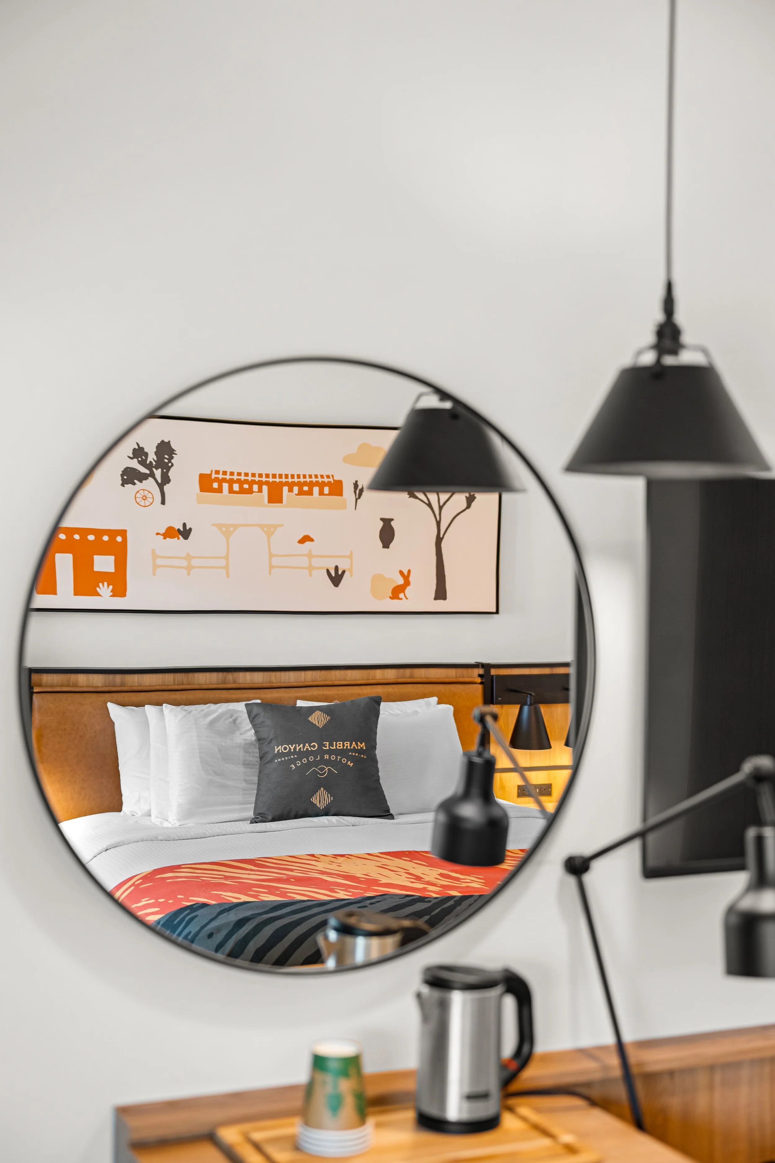 The photo shows a reflection of a neatly made bed with white pillows and a pillow with 'Marble Canyon Motor Lodge' on it, in a round mirror. There is a colorful bedspread, a wooden headboard, two black hanging lamps, and a framed artwork depicting a landscape scene with trees, a rabbit, and other animals.