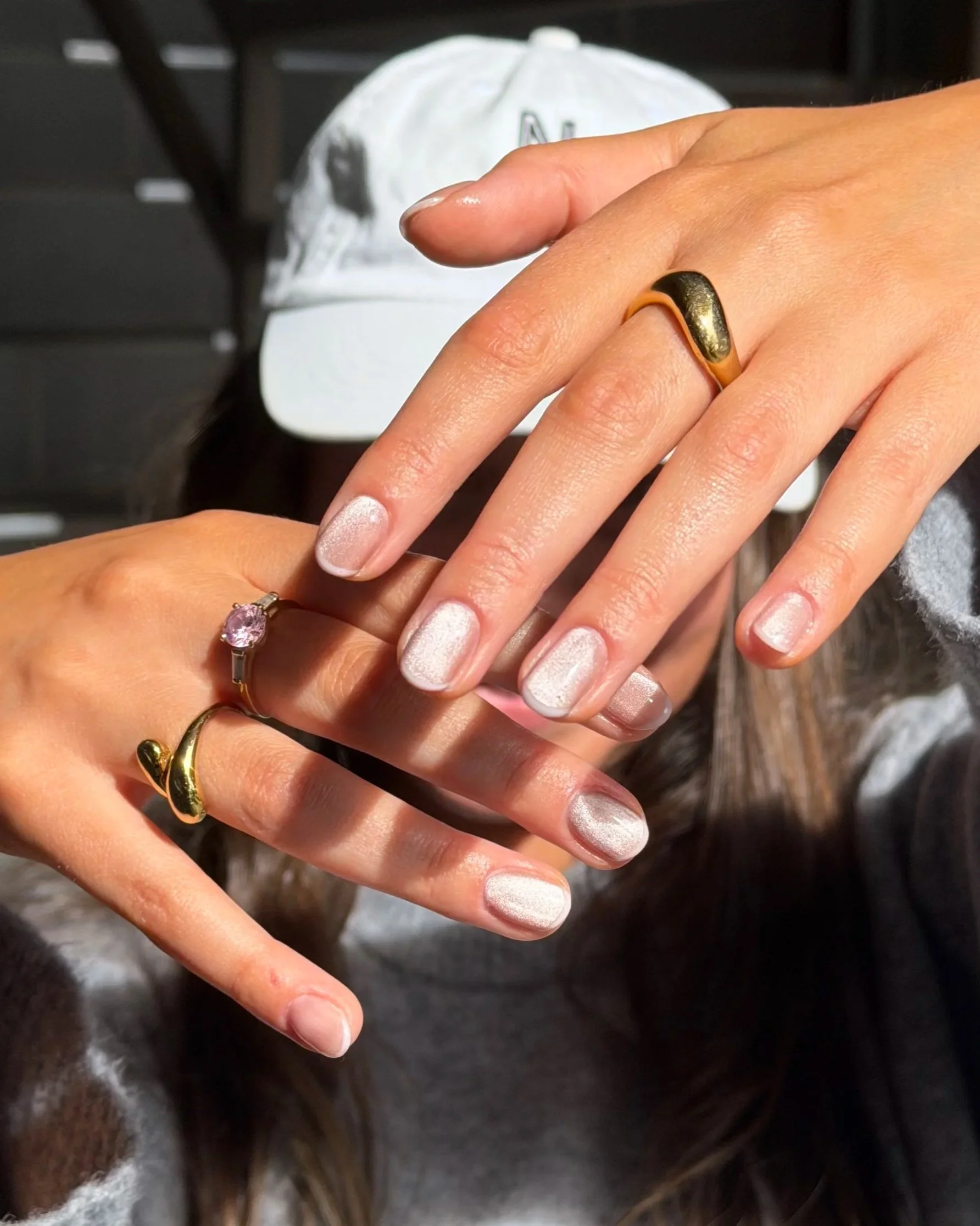 Close-up of two hands with fingers intertwined, wearing gold rings, one with a black stone ring and the other with a pink gemstone ring, with a gray cap in the background on a dark surface.