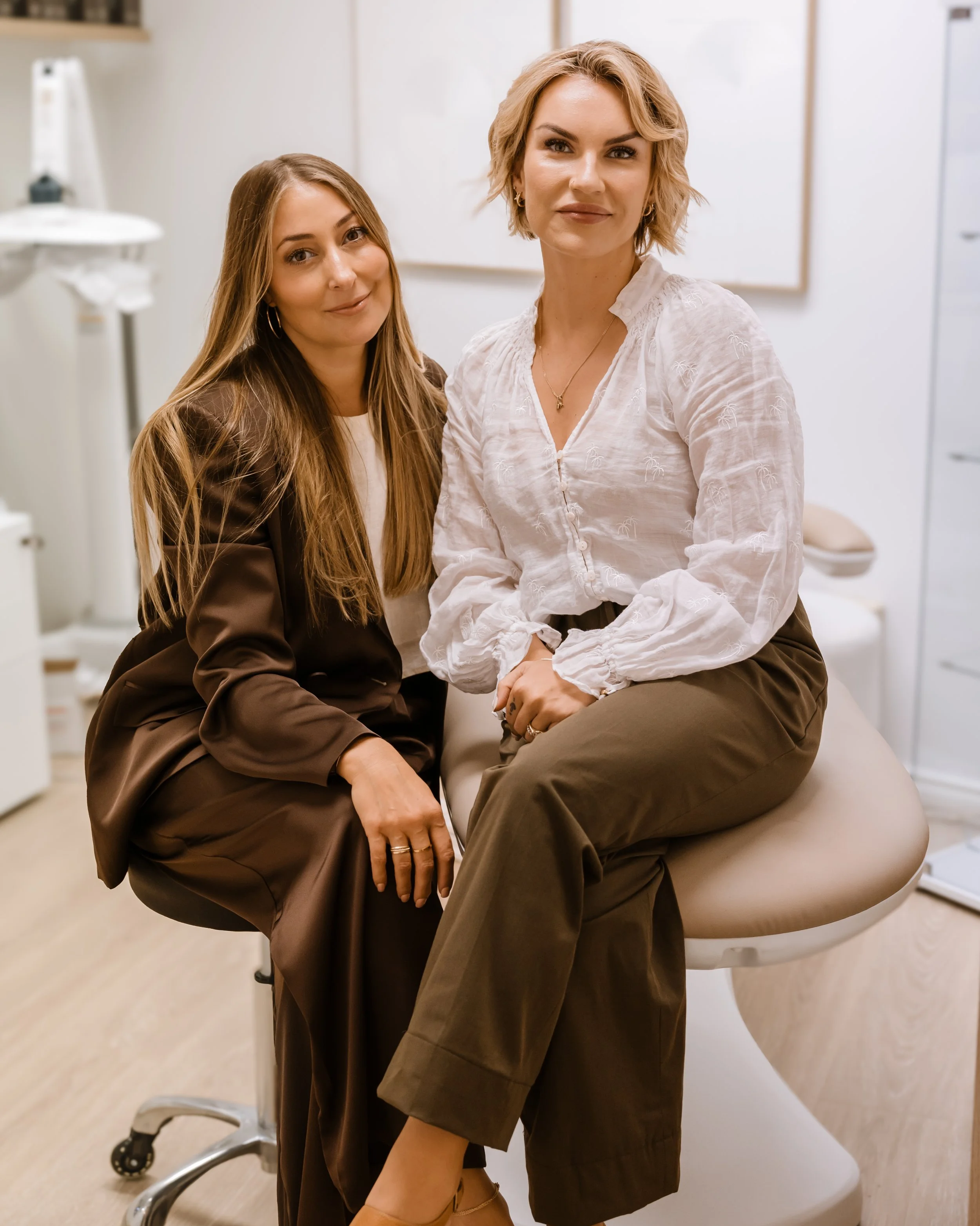 Two women sitting on a medical examination table in a clinic room, with one woman in a white shirt and the other in a brown outfit.