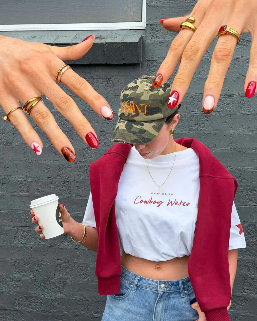A woman in casual clothing standing in front of a gray brick wall, holding a coffee cup in her left hand, wearing a white crop top with red text, light blue jeans, a red hoodie draped over her shoulders, and a camouflage cap. She is also wearing multiple rings, bracelets, and earrings.