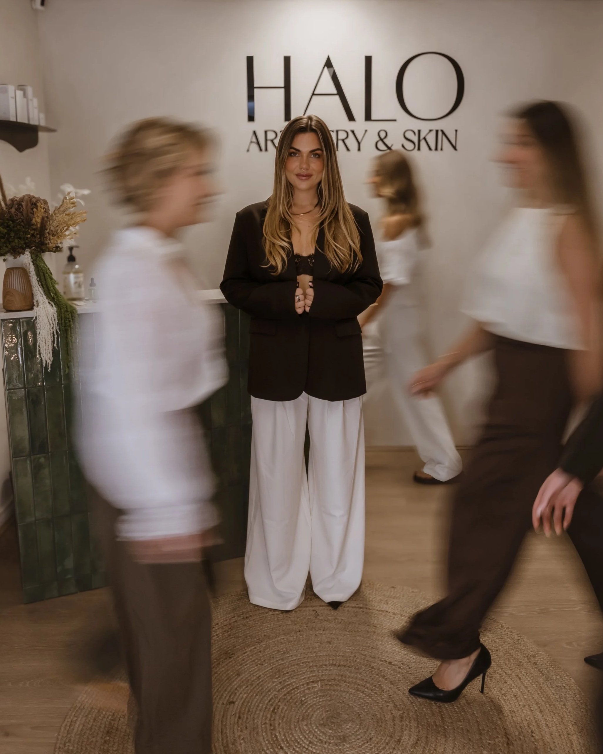 A woman with long brown hair and a black blazer standing inside a boutique or skincare store, with a sign behind her that reads 'HALO ARTISTRY & SKIN'. Several blurred figures of people walking by are visible in the background.