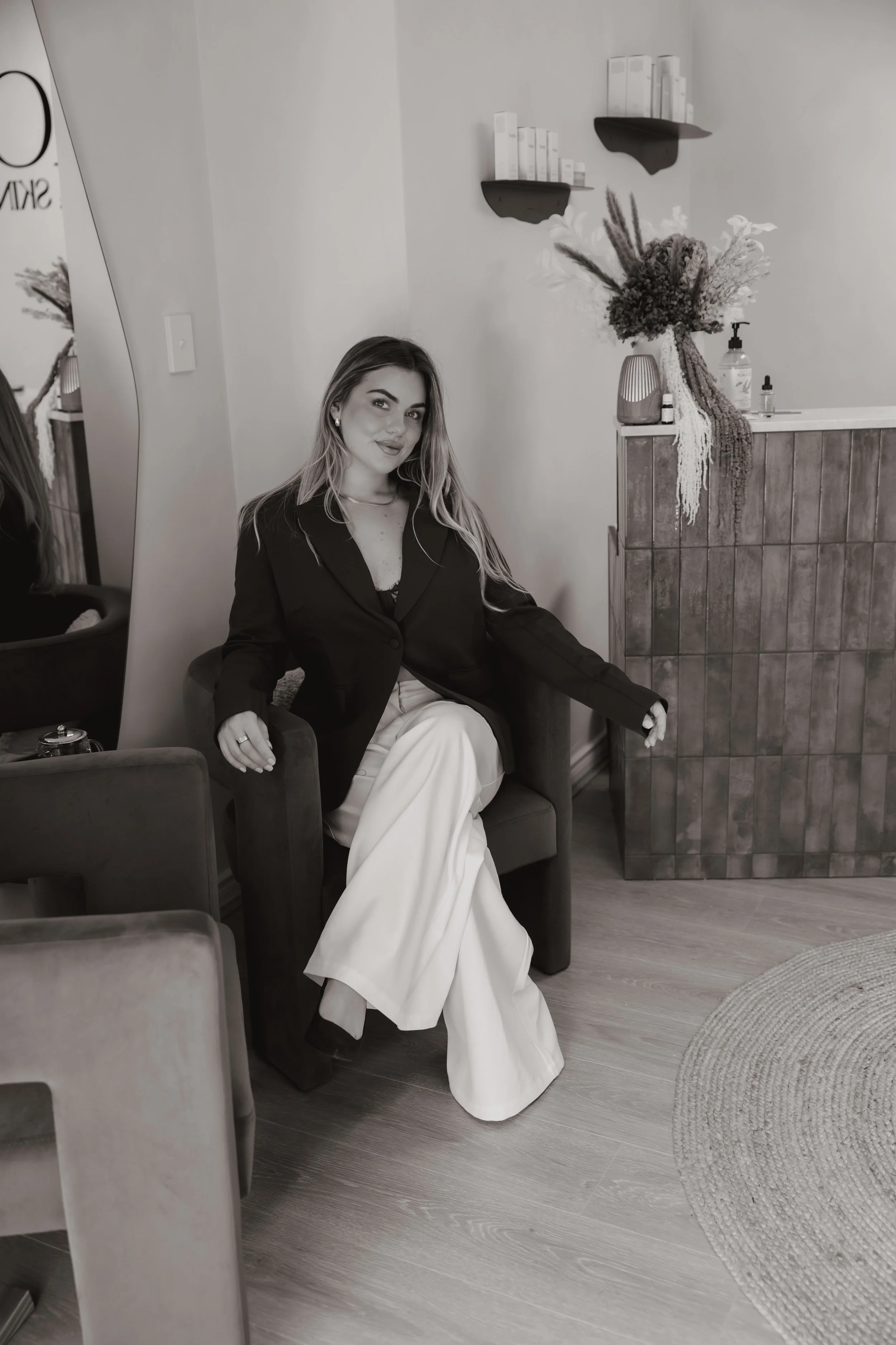 A woman with long hair and earrings sitting in a chair, wearing a dark blazer and light-colored pants, in a room with a vase of flowers, a mirror, bookshelf, and a wooden panel on the wall.