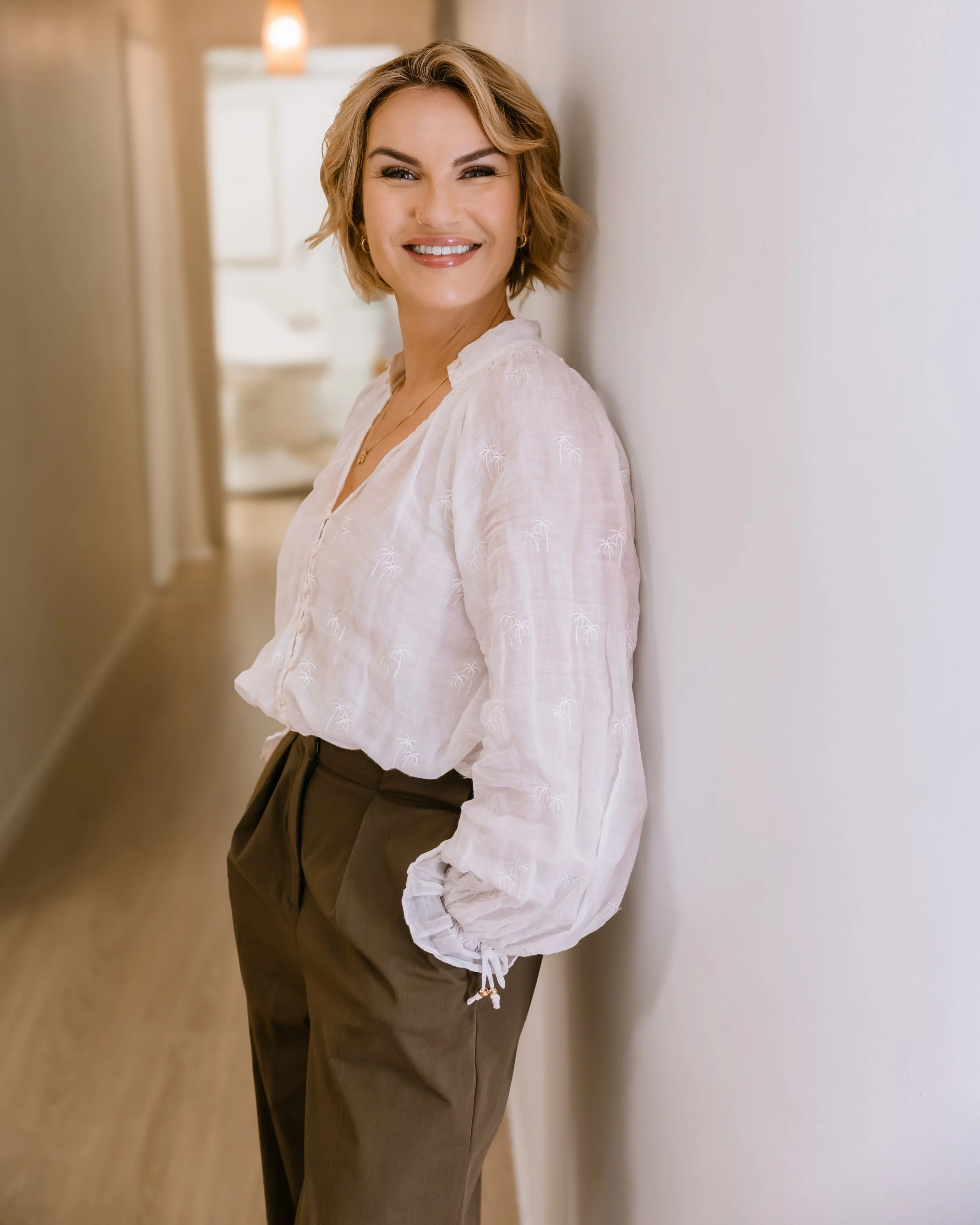 A smiling woman with short blonde hair, wearing a white blouse with small palm tree print and brown pants, leaning against a white wall in a warm, softly lit indoor setting.