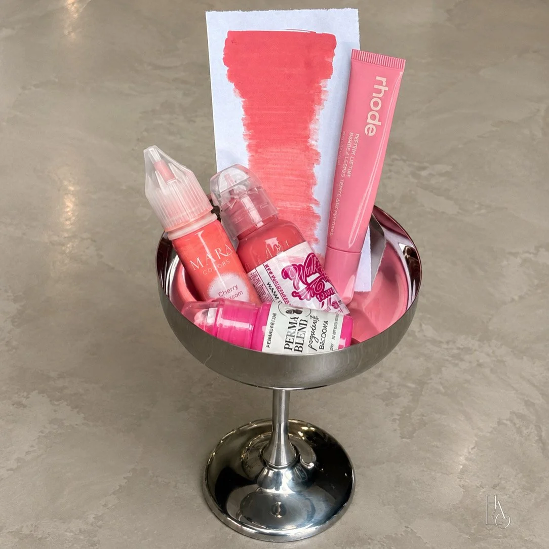 A metal bowl on a stand filled with various pink skincare and cosmetic products, including a tube, bottles, and a piece of paper with a pink swatch.